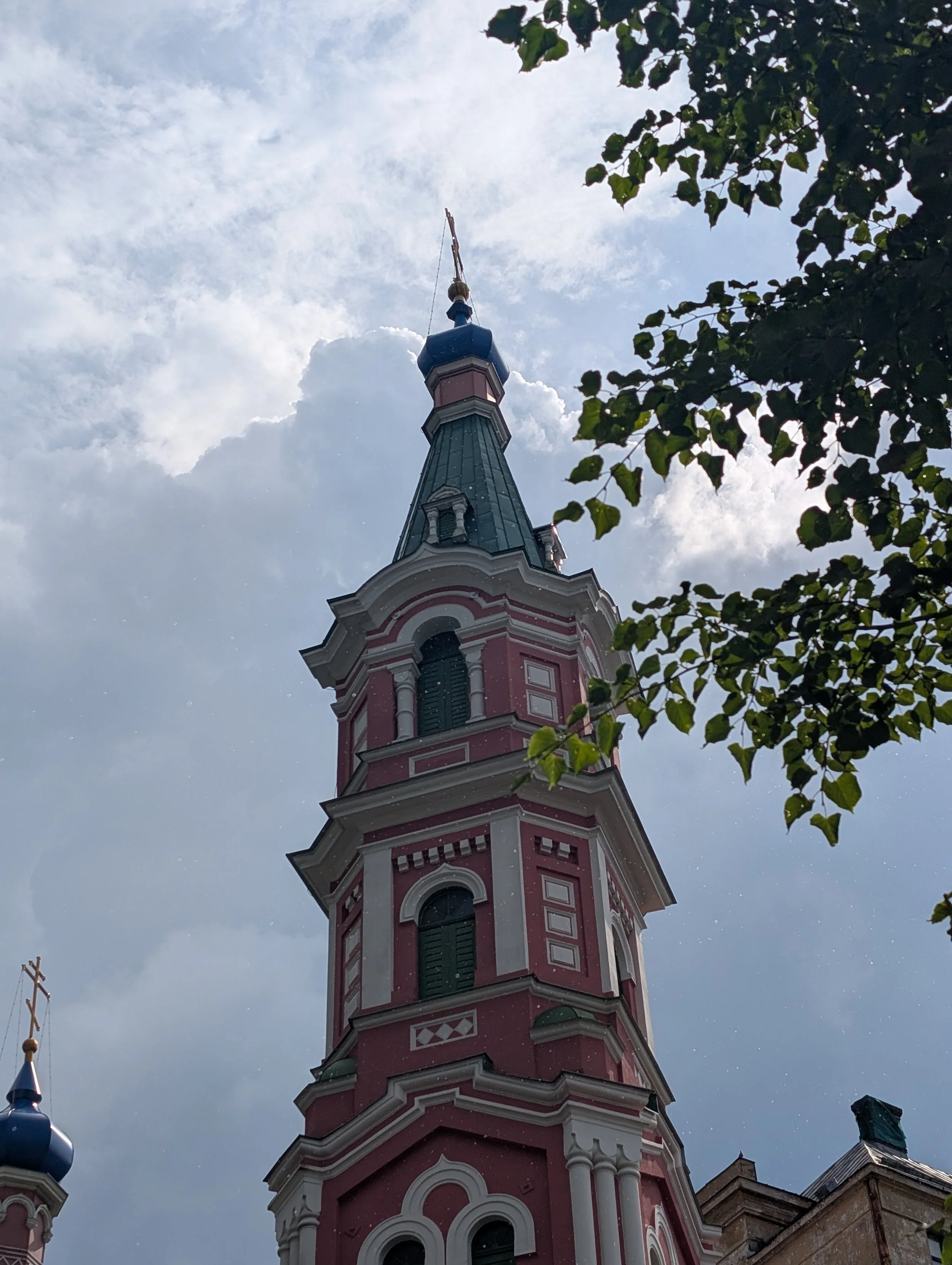 View of a tall, pink and white church tower with a blue spire, set against a cloudy sky, partially obscured by green tree leaves.
