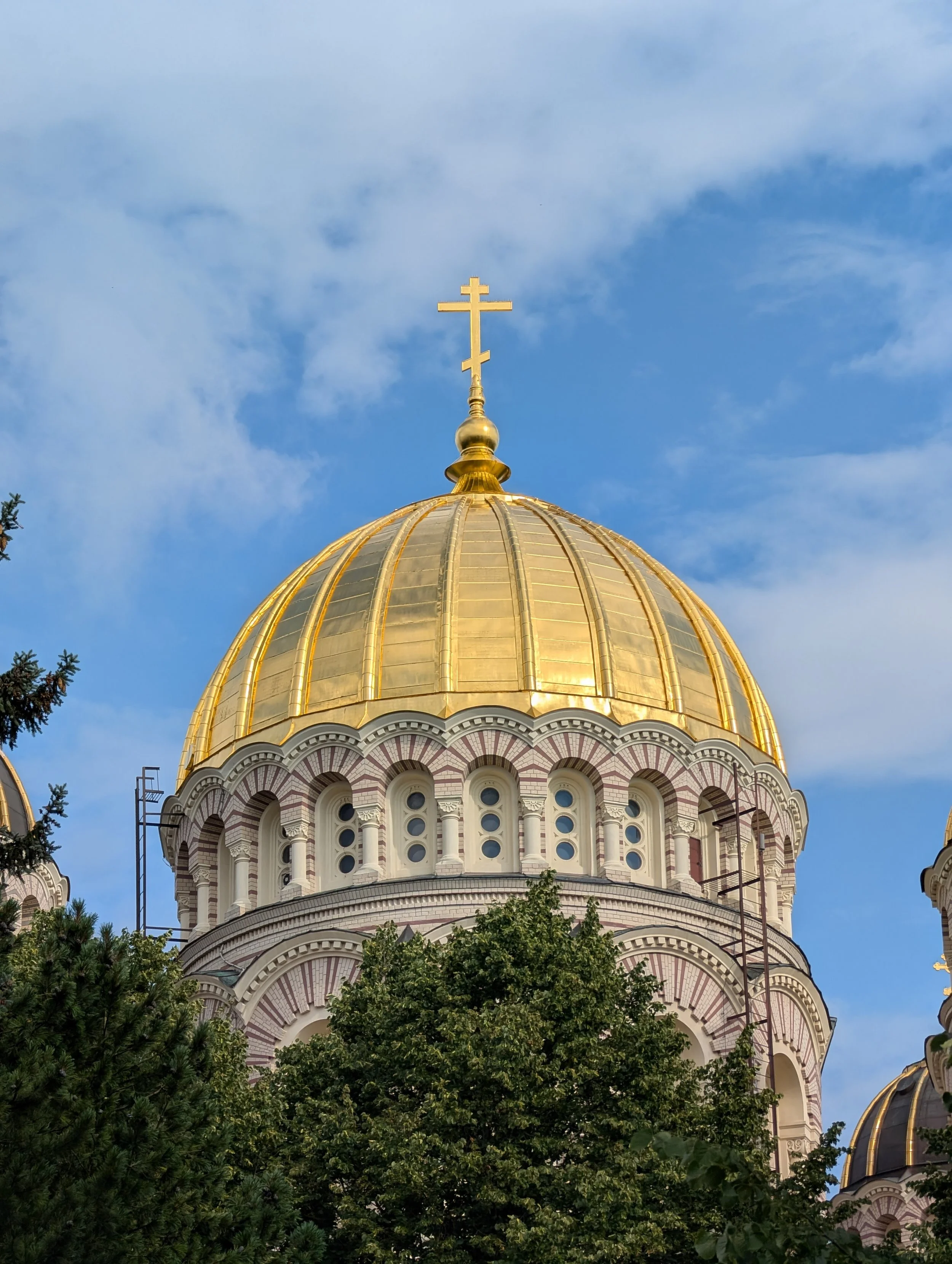 A large golden dome with a cross on top, part of an Orthodox church, surrounded by trees and set against a blue sky with light clouds.