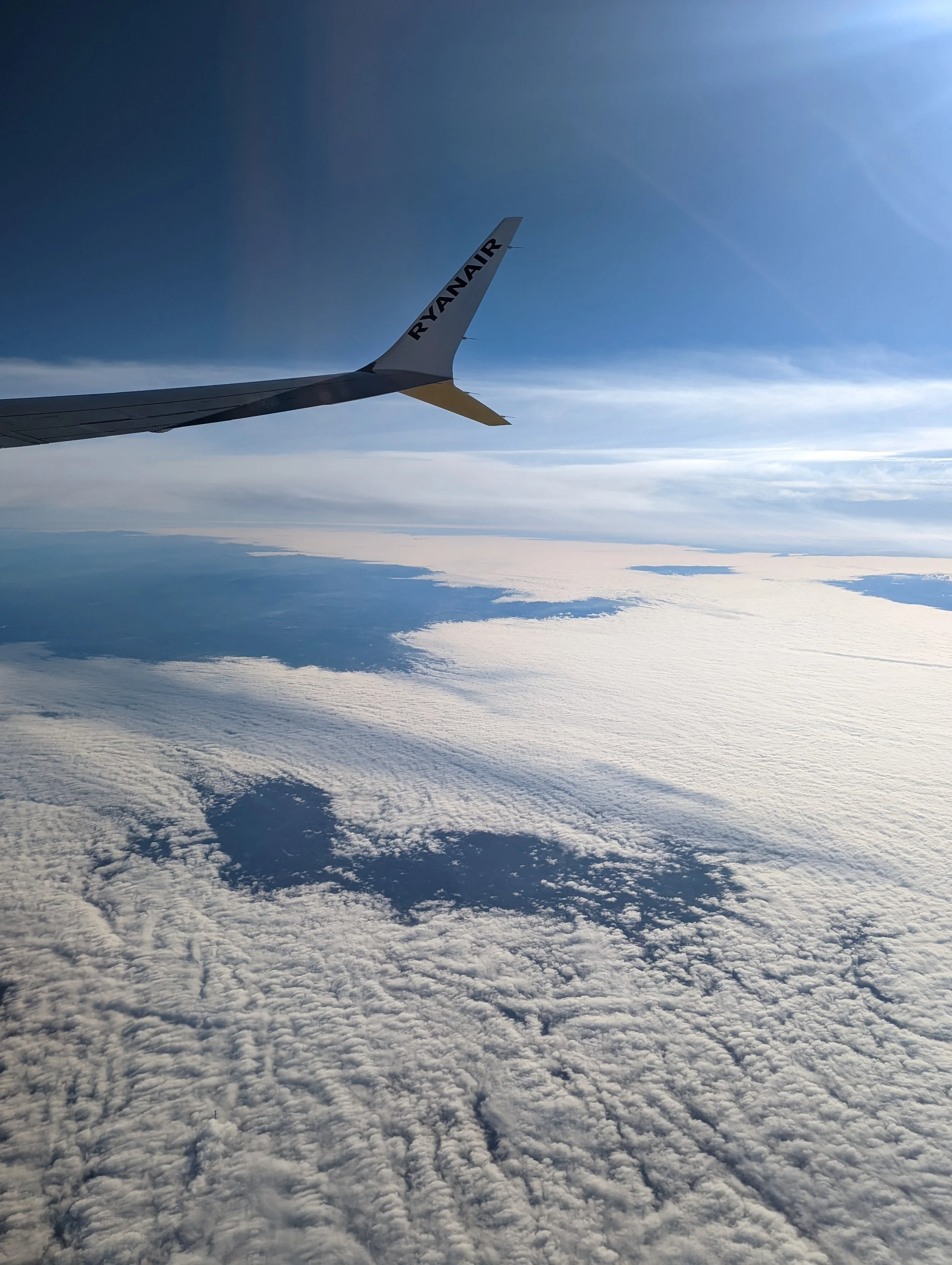 View from an airplane window showing aircraft wing with 'RYANAIR' branding and a vast cloud-covered sky below.