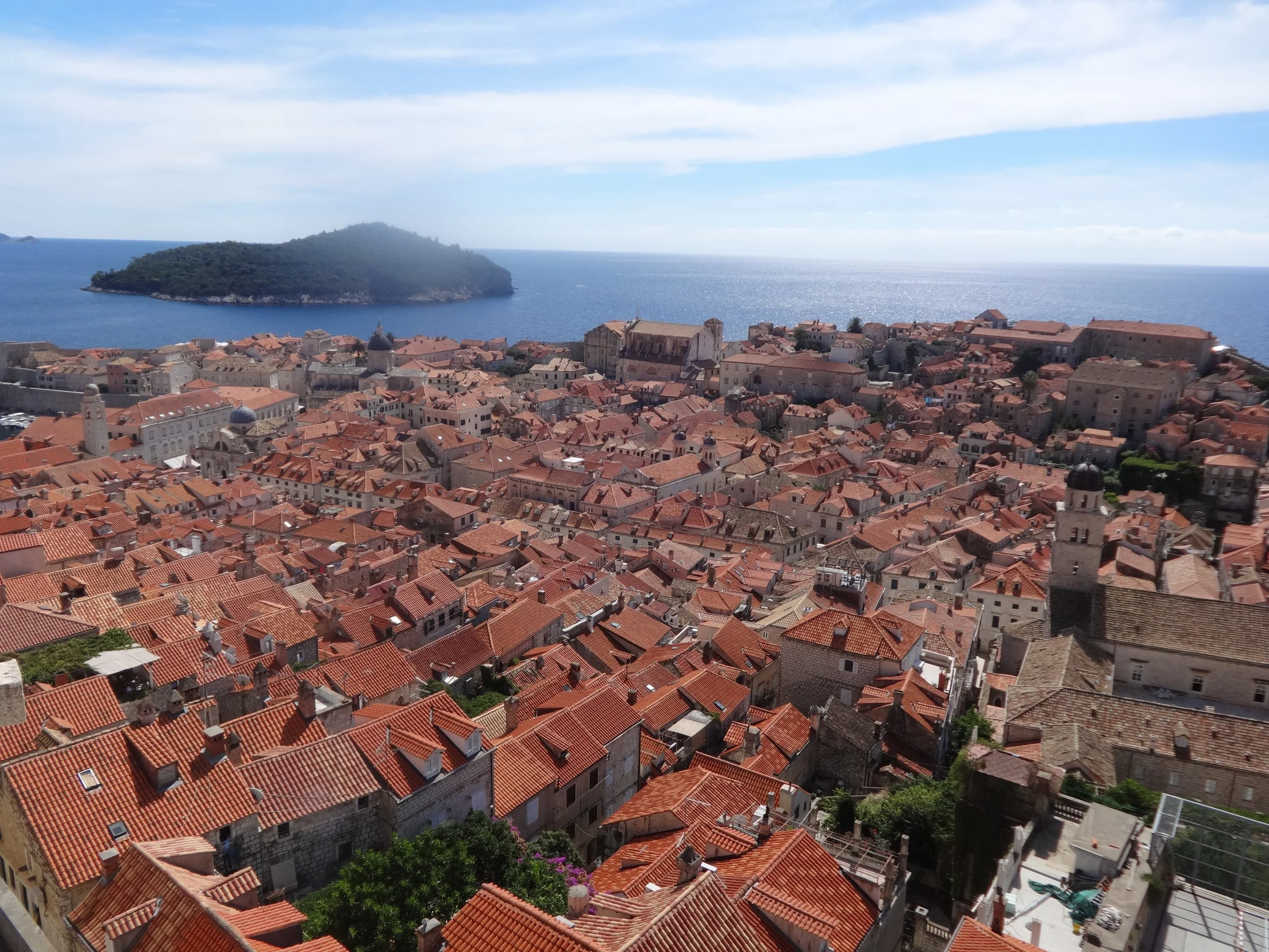A coastal town with a dense cluster of buildings with red-tiled roofs, overlooking the ocean with a small island in the distance, and a partly cloudy blue sky.
