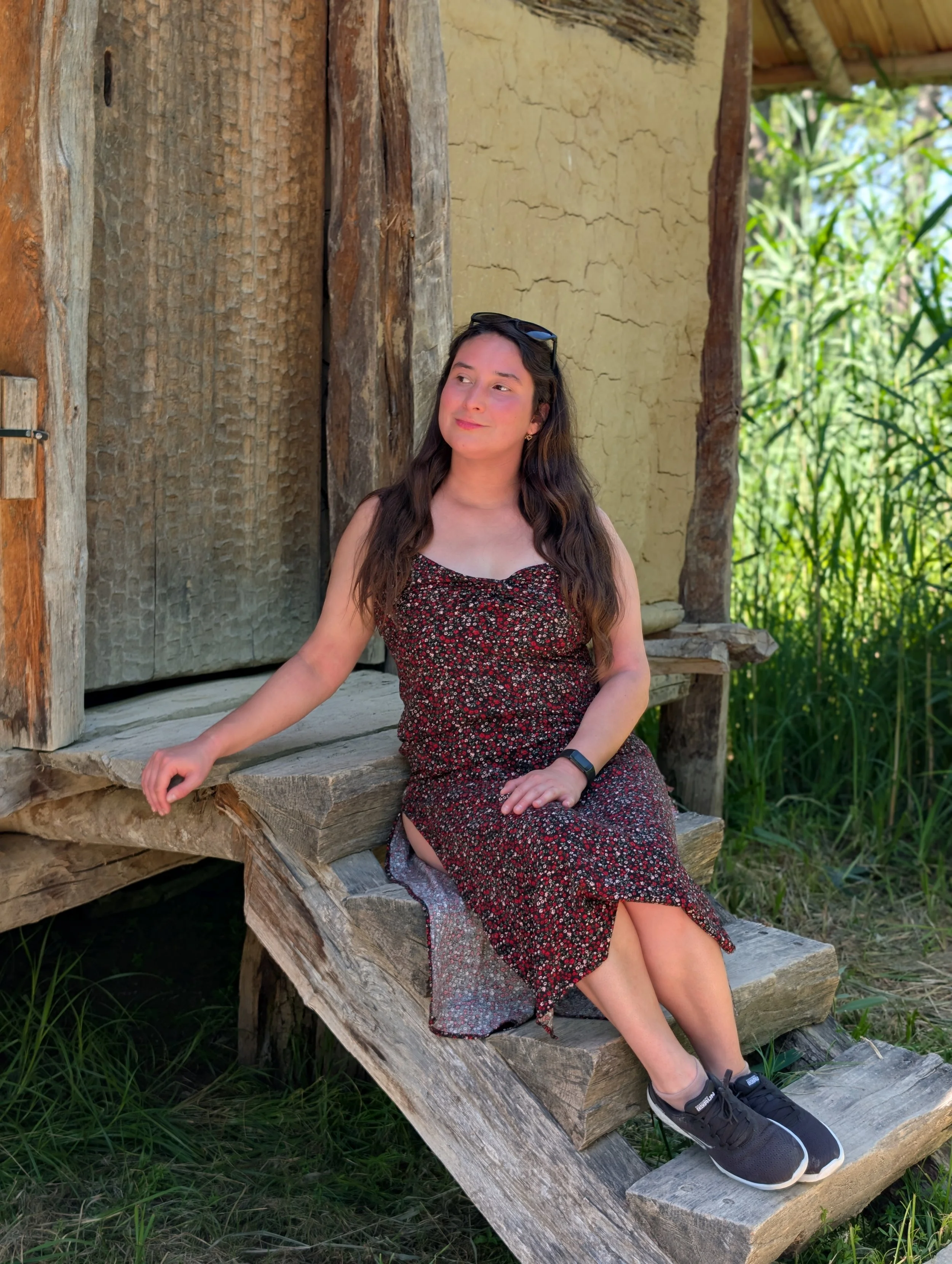A young woman with long dark hair sitting on wooden stairs outside a rustic building with a yellow wall, green foliage in the background, wearing a patterned dress, black sneakers, and sunglasses resting on her head.
