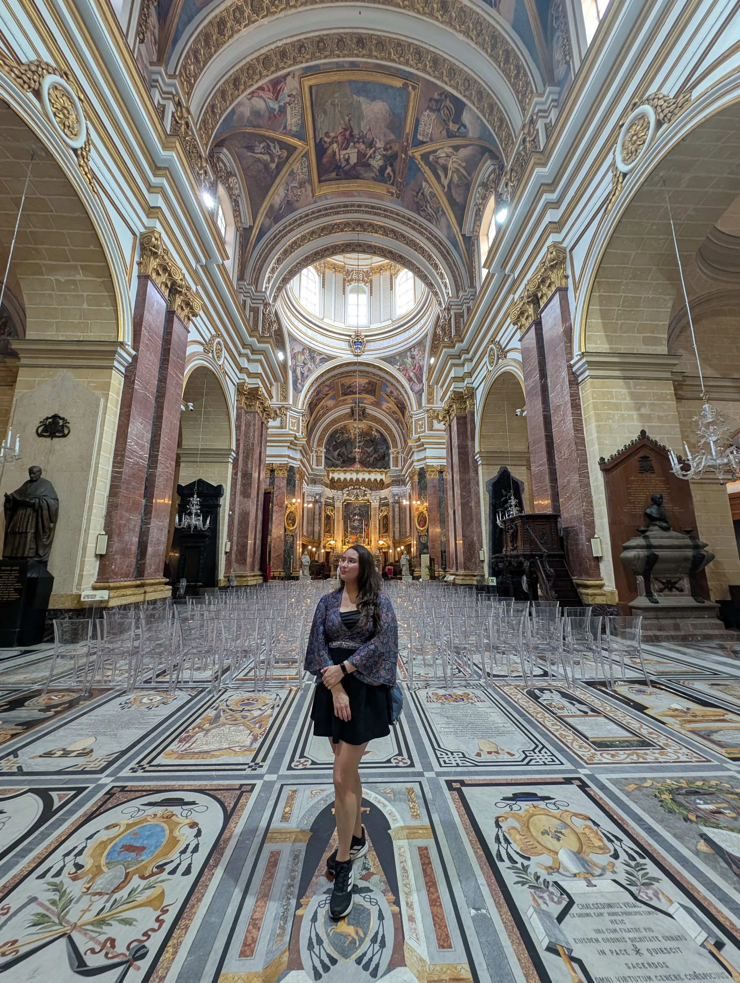 Inside a grand, ornate church with decorated arches, paintings, and statues. A woman stands in front, wearing a dark dress and high-top sneakers, amidst a detailed patterned floor with various coats of arms and inscriptions.