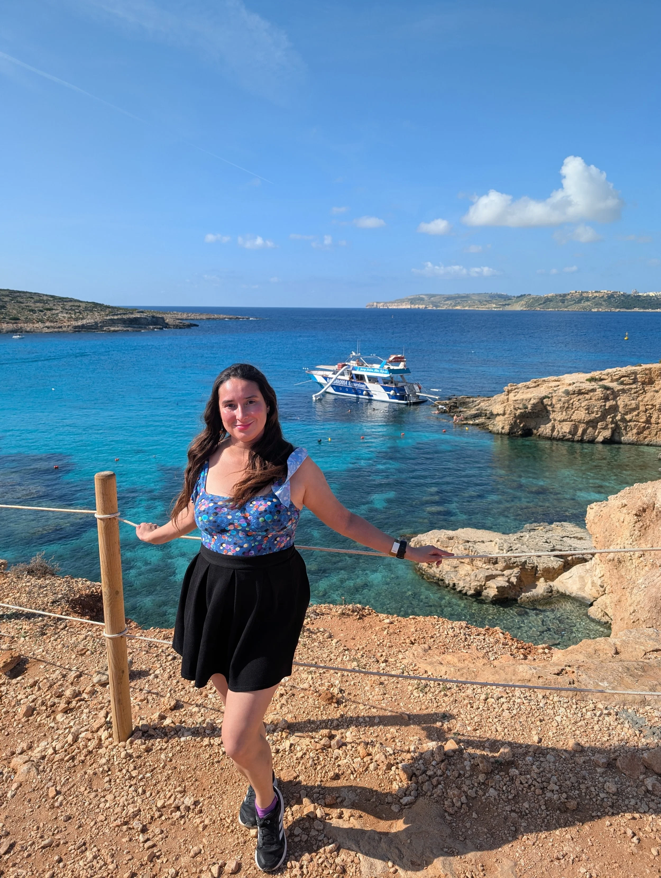 A woman with long dark hair wearing a blue floral top and a black skirt standing on rocky ground near a railing, with clear blue water, boats, rocks, and a distant coastline with buildings under a partly cloudy sky in the background.