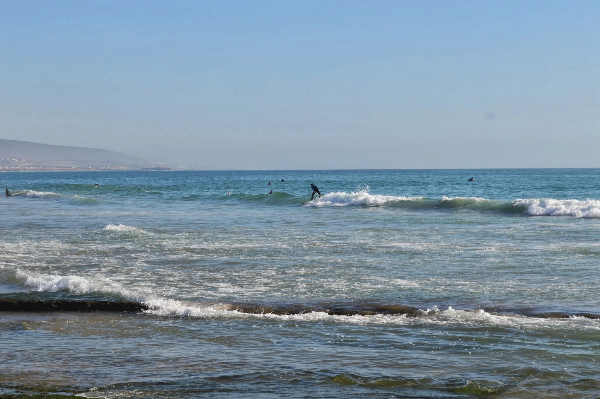 People surfing and swimming in the ocean near a sandy beach with a distant coastline and hills under a clear blue sky.