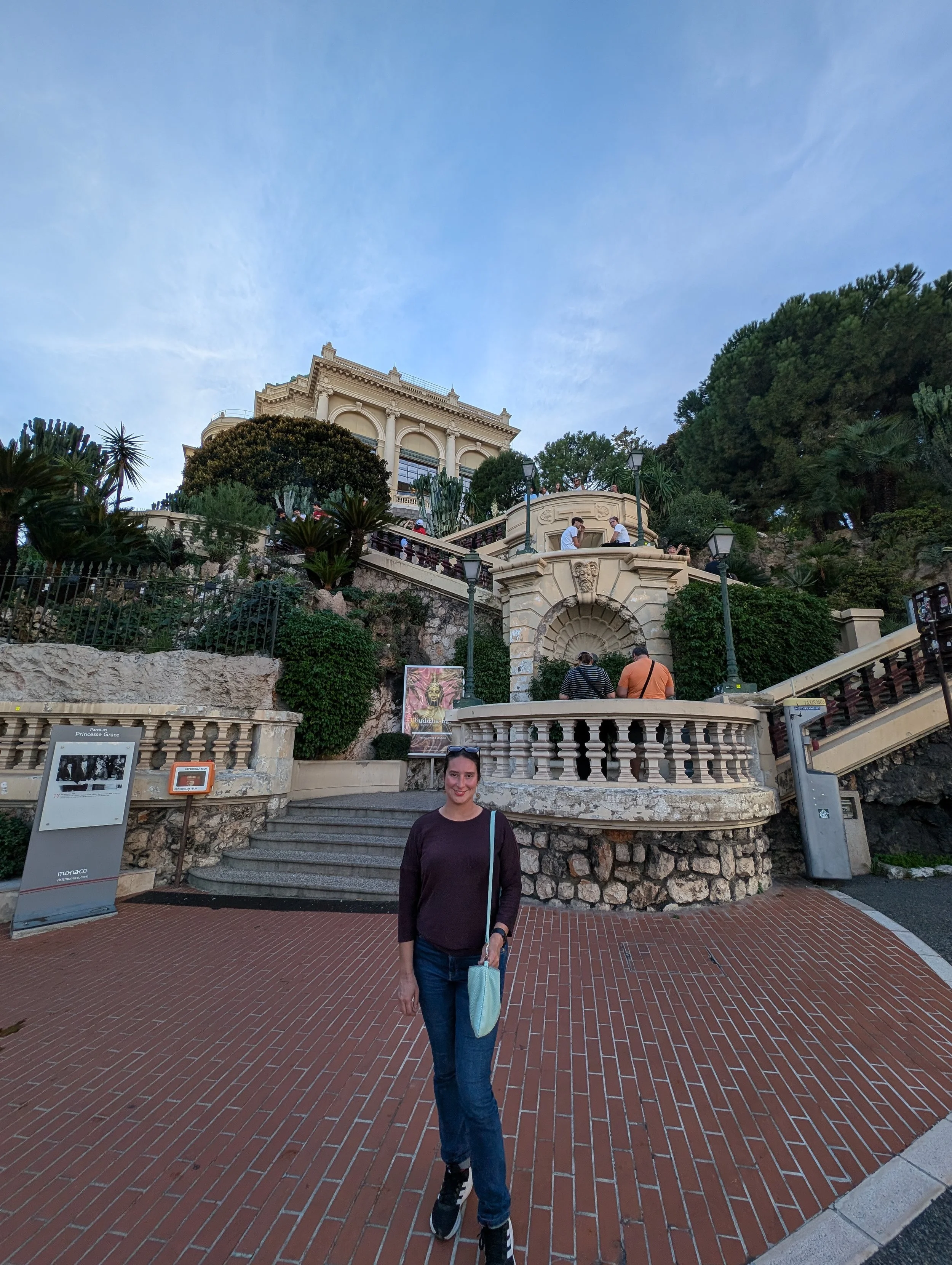 A woman stands on a brick-paved area in front of a ornate historic building and lush greenery, with a smiling expression and casual clothing.
