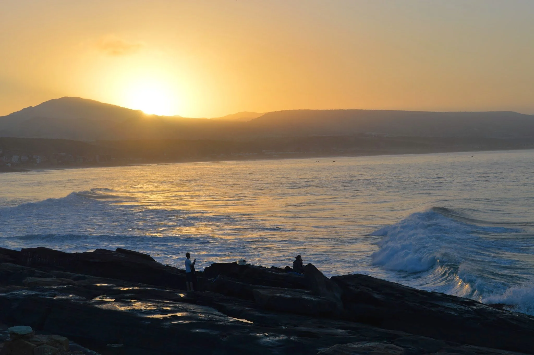 Sunset over the ocean with waves crashing against rocks along the shoreline; two people standing on rocks watching the sunset, with hills in the background.