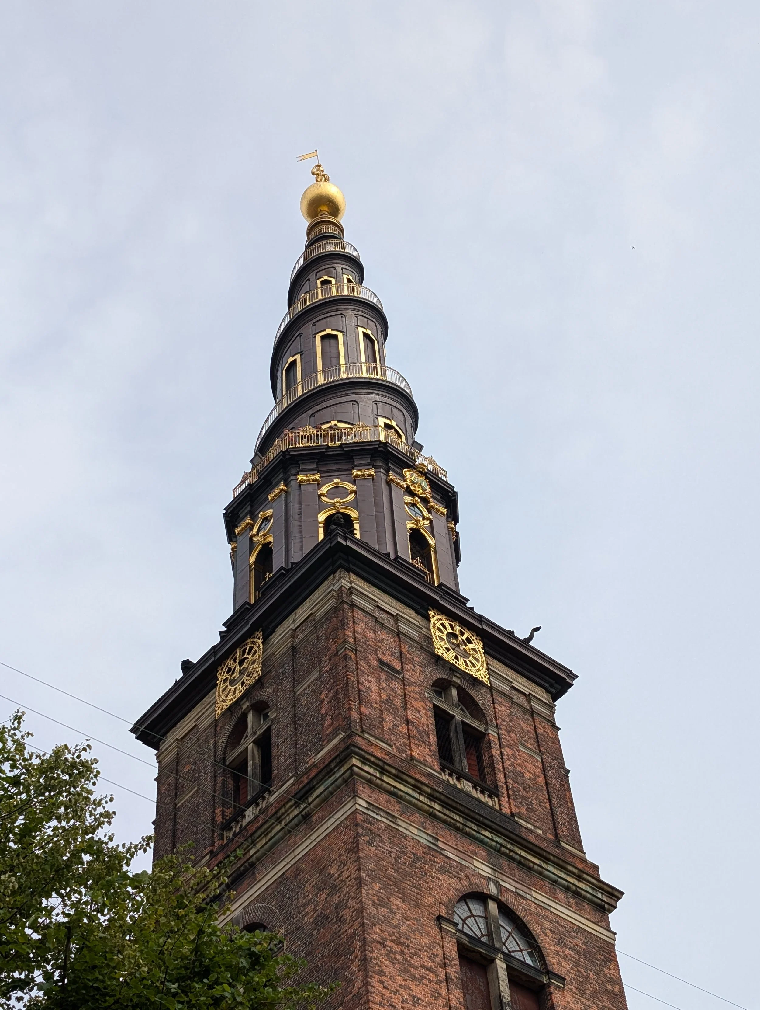 A tall brick clock tower with gold accents and a gold sphere at the top, under a cloudy sky, with some tree foliage in the foreground.