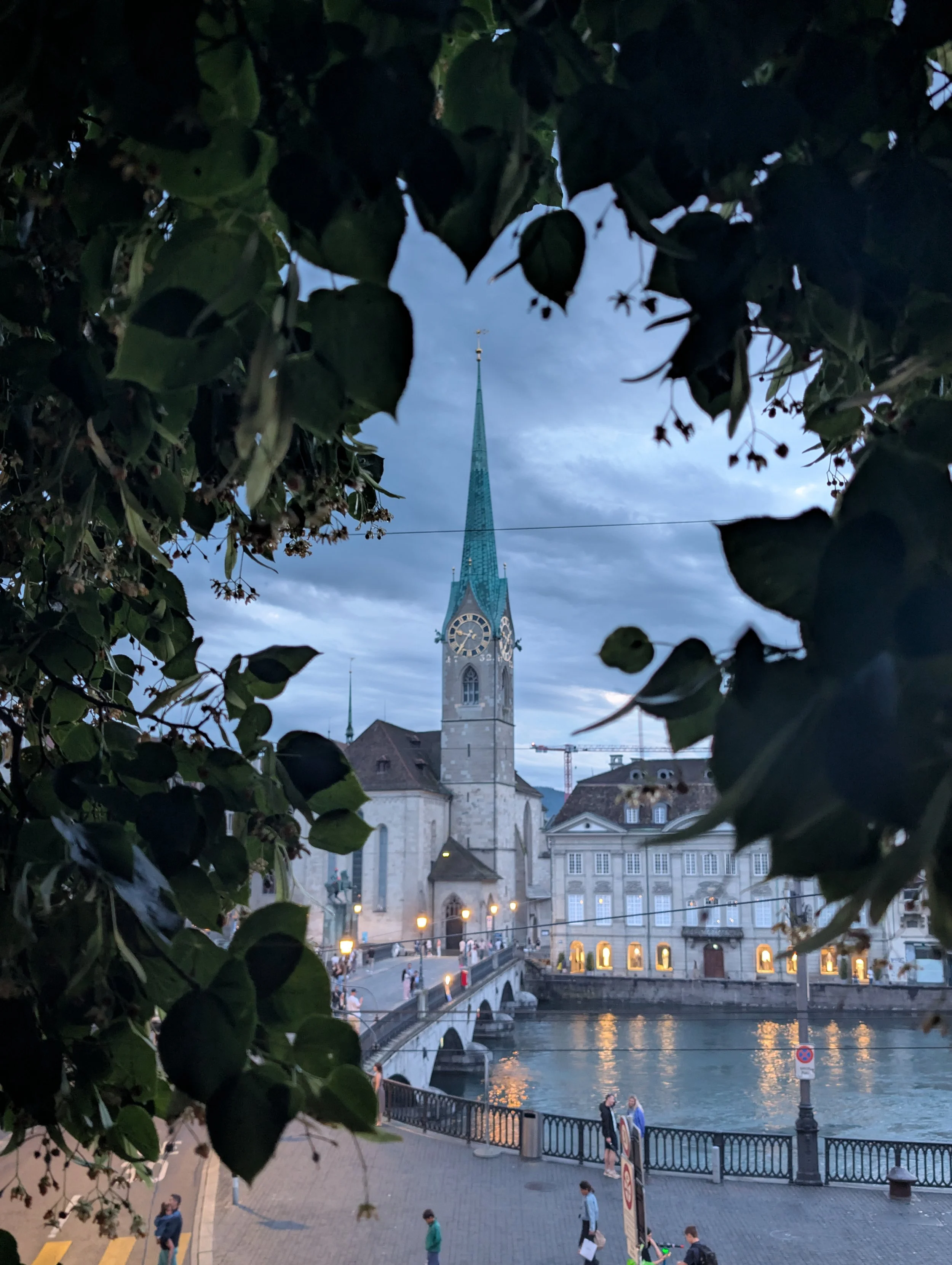 City square with a river, a stone bridge, and buildings in the background. A church with a tall spire and clock is visible, with leaves framing the scene. People are walking and gathering along the riverbank in the evening.