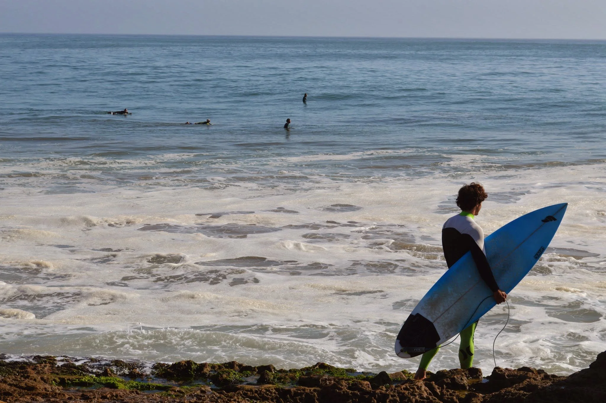 A woman in a wetsuit holding a surfboard walking along the rocky shoreline of the ocean with waves and four surfers in the water.