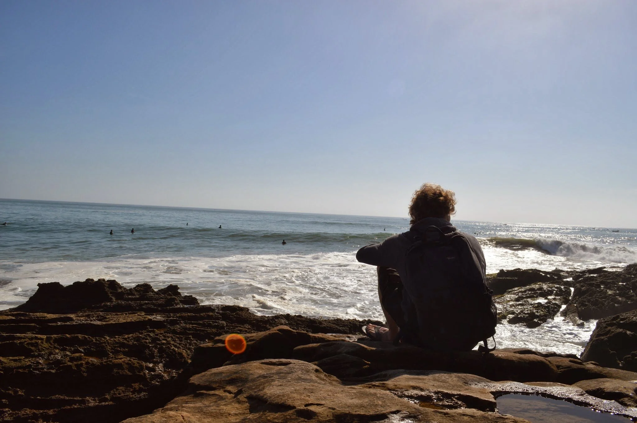 A person with curly hair sitting on rocks by the beach, overlooking the ocean with some surfers in the water, under a clear sky.