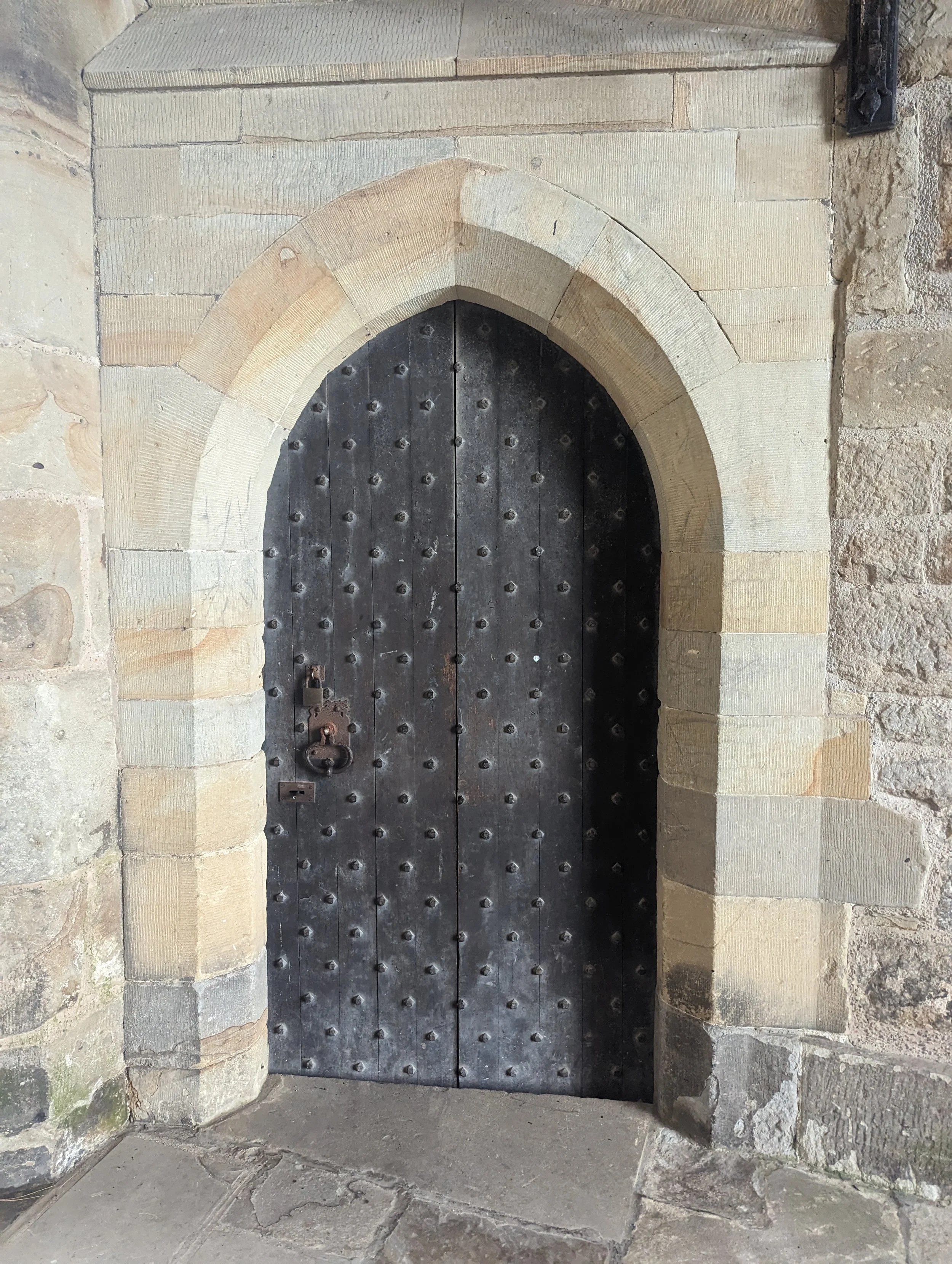 A black, metal door with rivets, set within a stone archway made of beige and gray stones.