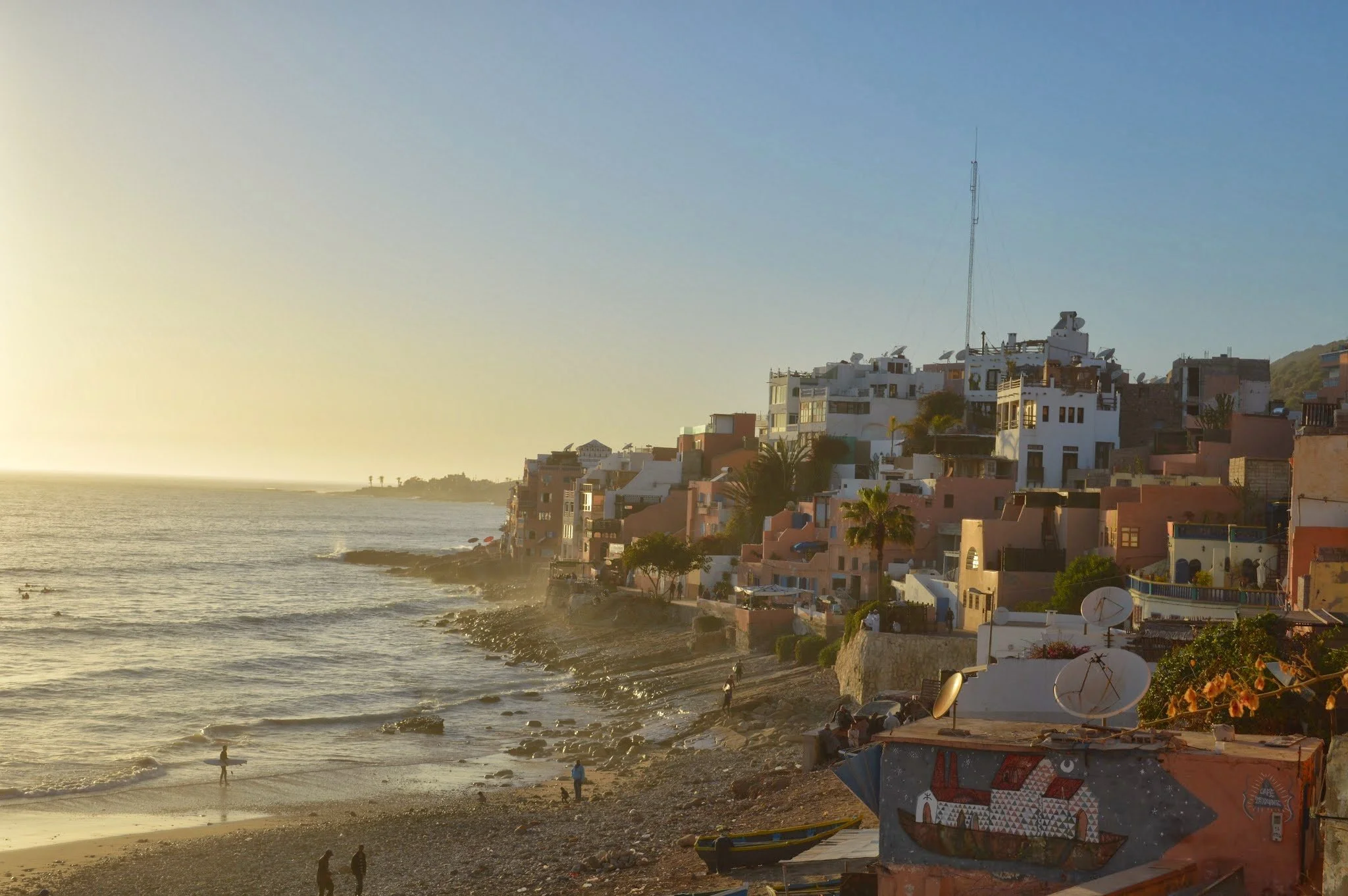 A coastal cityscape during sunset with buildings on a hillside overlooking the ocean and people walking along the rocky beach.