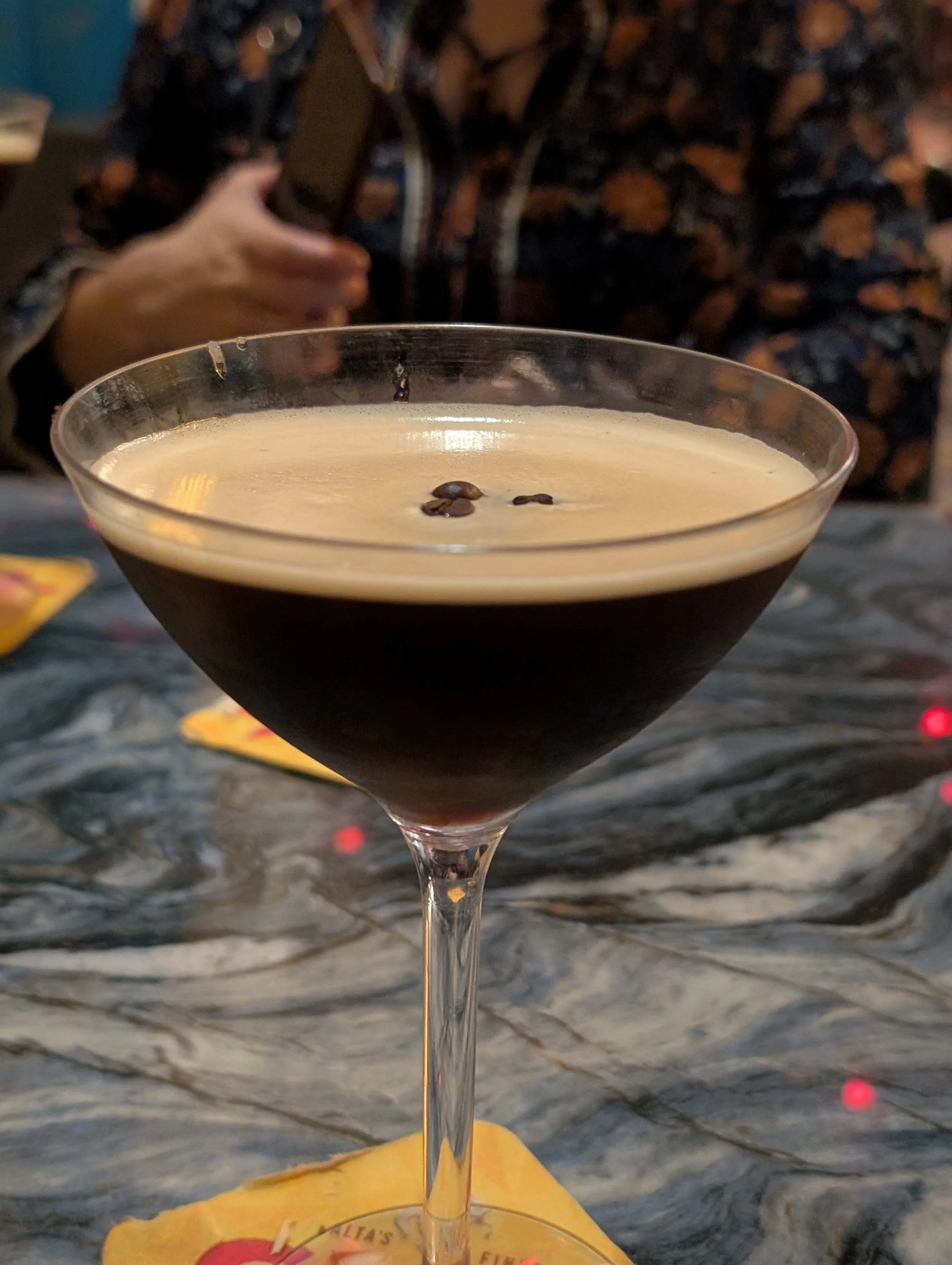 A glass of coffee with coffee beans on top, placed on a table with a yellow napkin underneath. In the background, a person wearing a dark, patterned shirt is reaching for or holding something, out of focus.