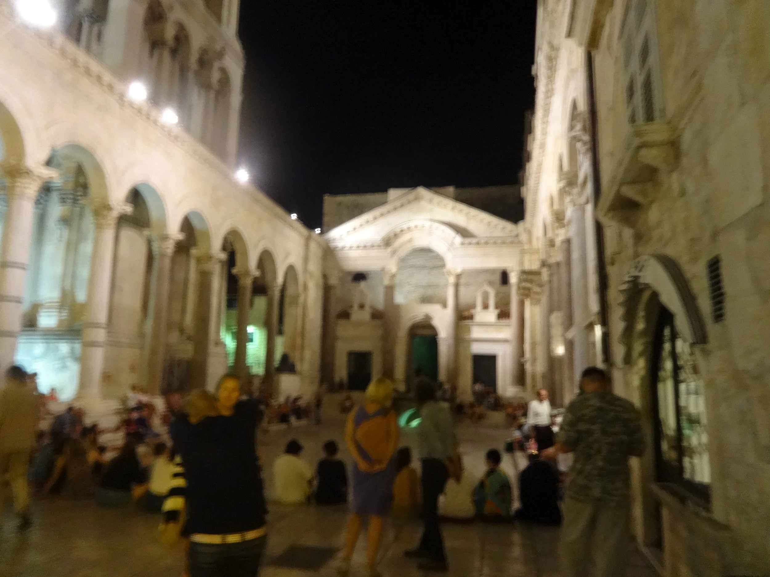 Nighttime scene inside a large, classical-style church or cathedral with ornate architecture, arches, and columns, populated with people sitting and standing, some wearing yellow and black uniforms.