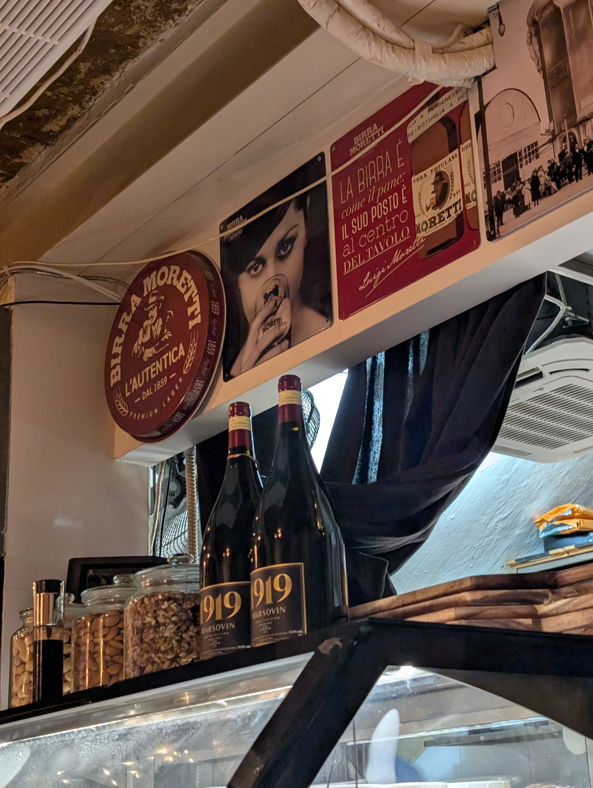 Shelf with bottles of wine, jars of nuts, and various signs and posters, including vintage beer advertisements. The image is taken from inside a cafe or shop.