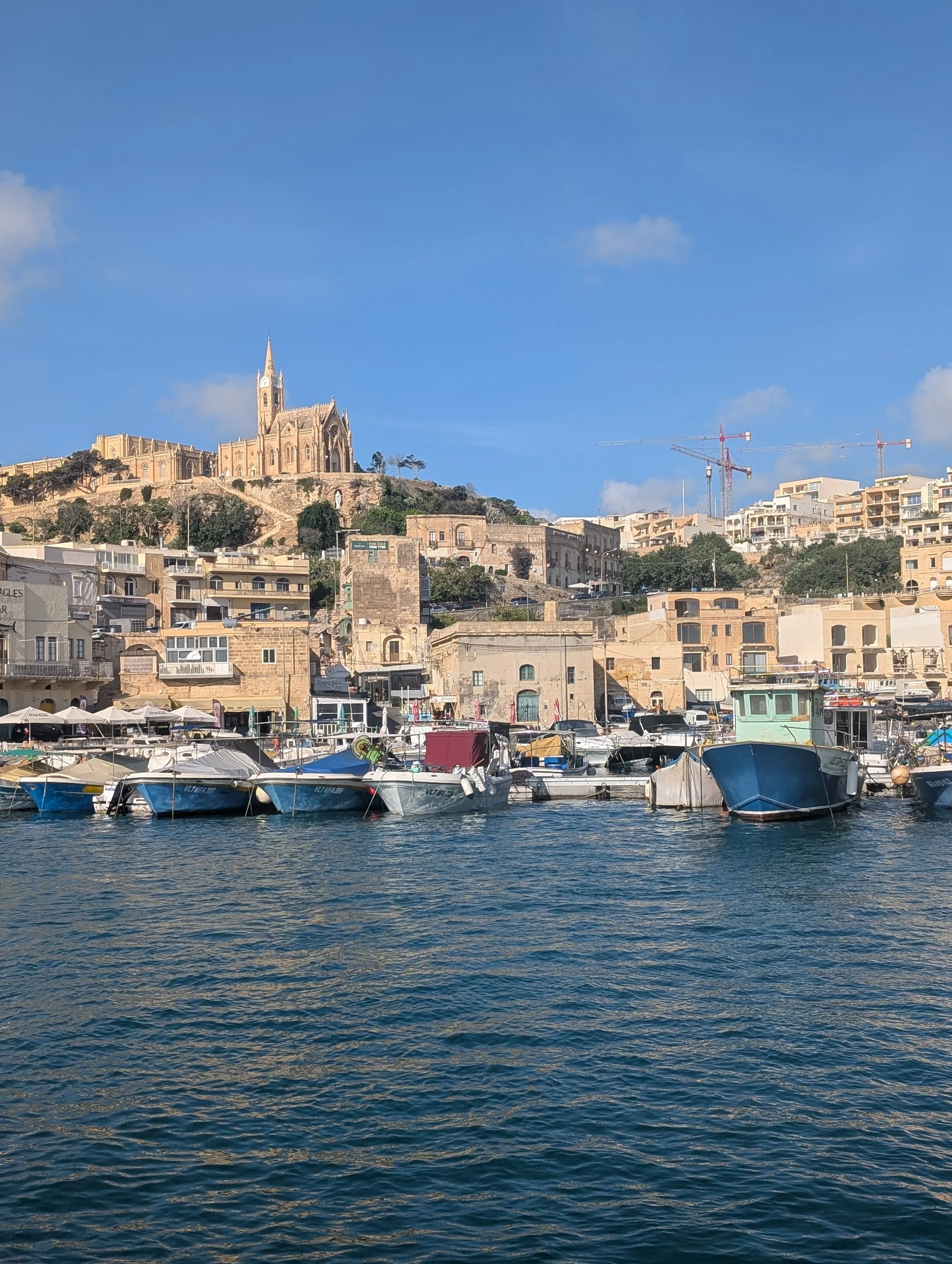 Boats docked at a harbor with buildings and a church on a hill in the background under a cloudy blue sky.