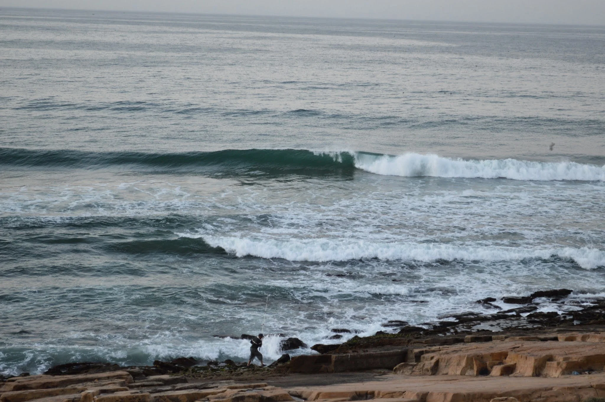 People fishing by rocky shoreline with ocean waves in the background.