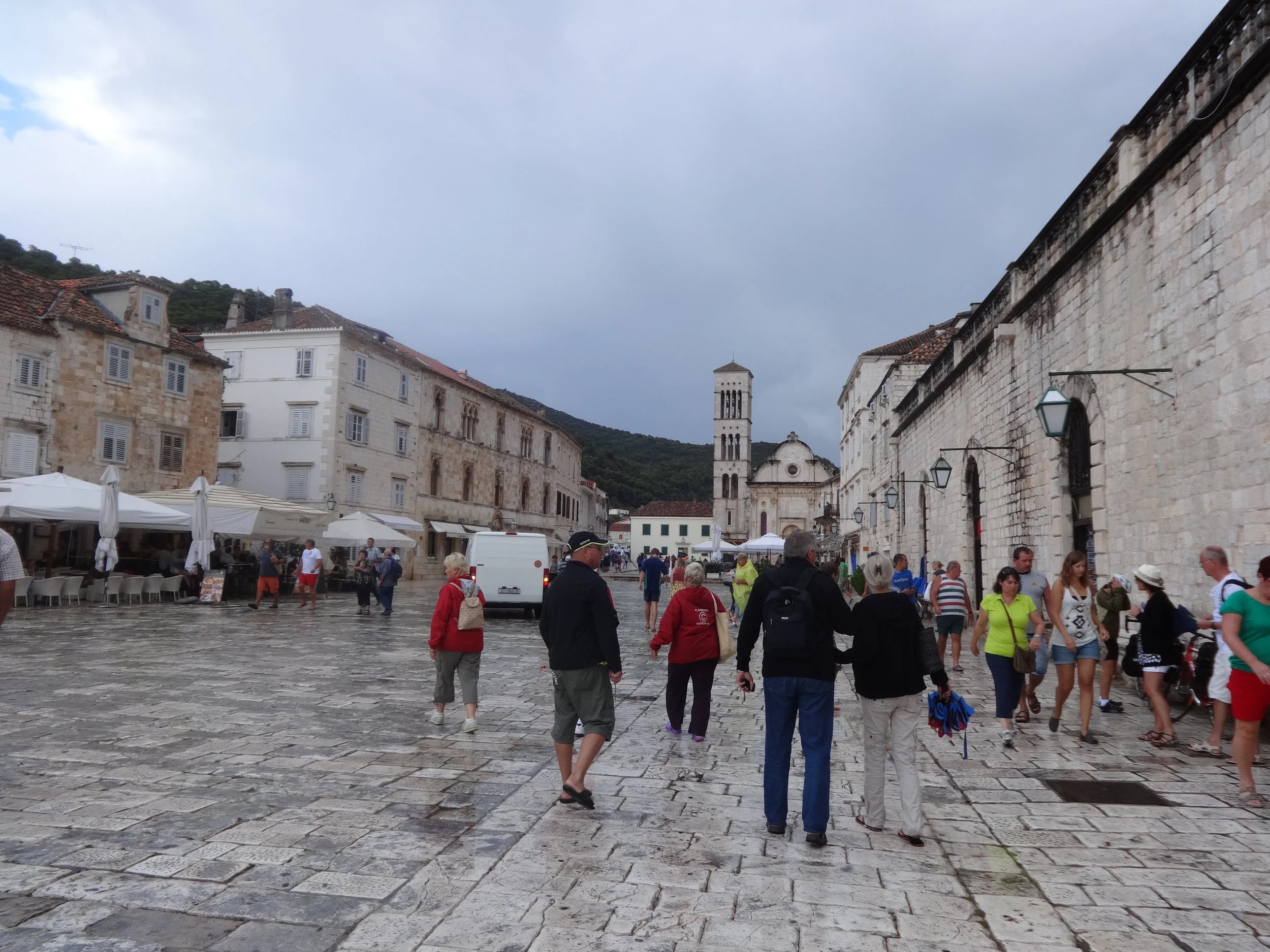 Crowd of tourists walking on cobblestone square in historic European city, with old stone buildings, outdoor cafes, and church tower under cloudy sky.