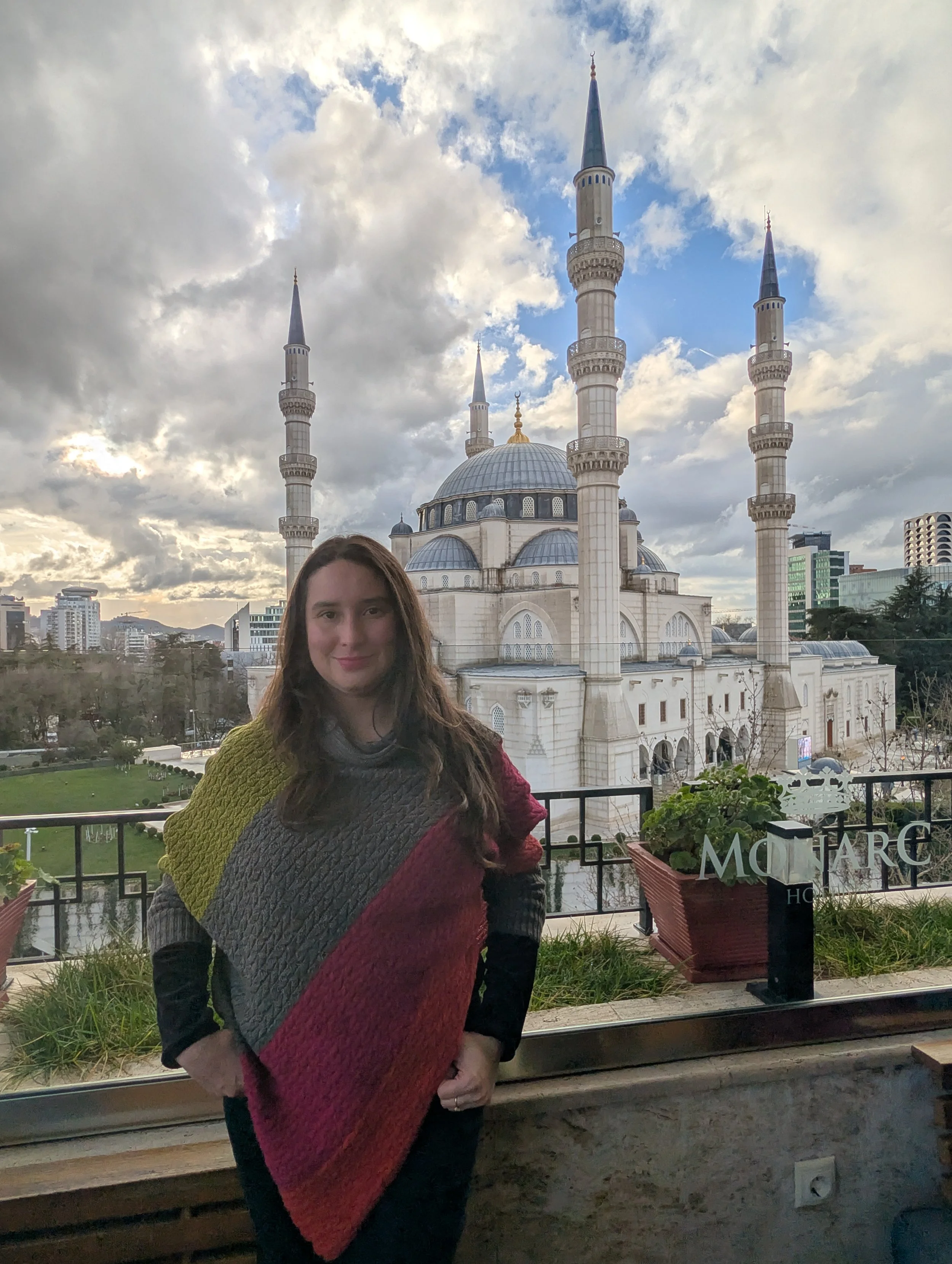 Tamara with long brown hair wearing a multicoloured sweater standing on a balcony with a cityscape and a mosque with minarets and a dome in the background.
