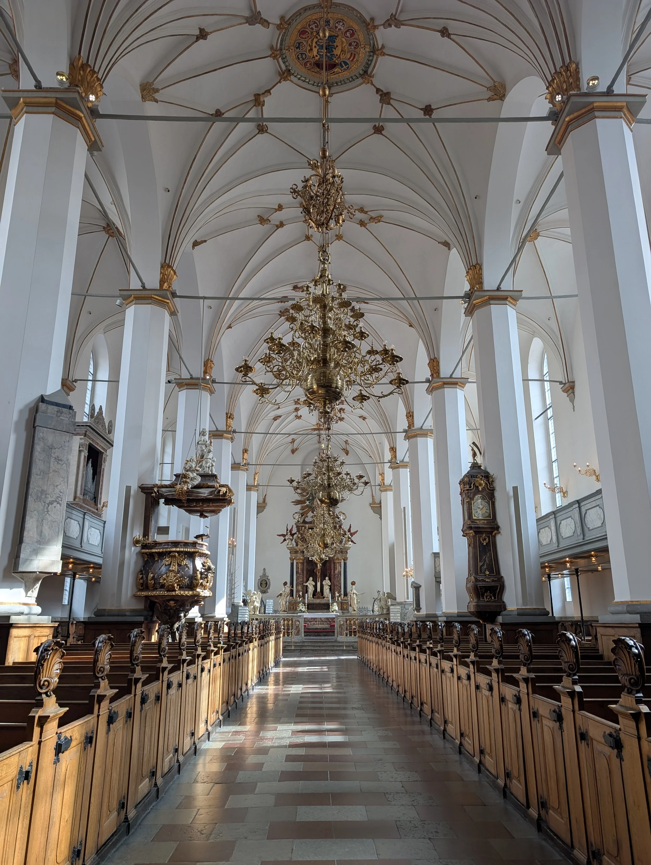 Interior of a grand church with white walls, tall windows, ornate golden chandeliers, and wooden pews arranged in rows facing an altar with religious statues.