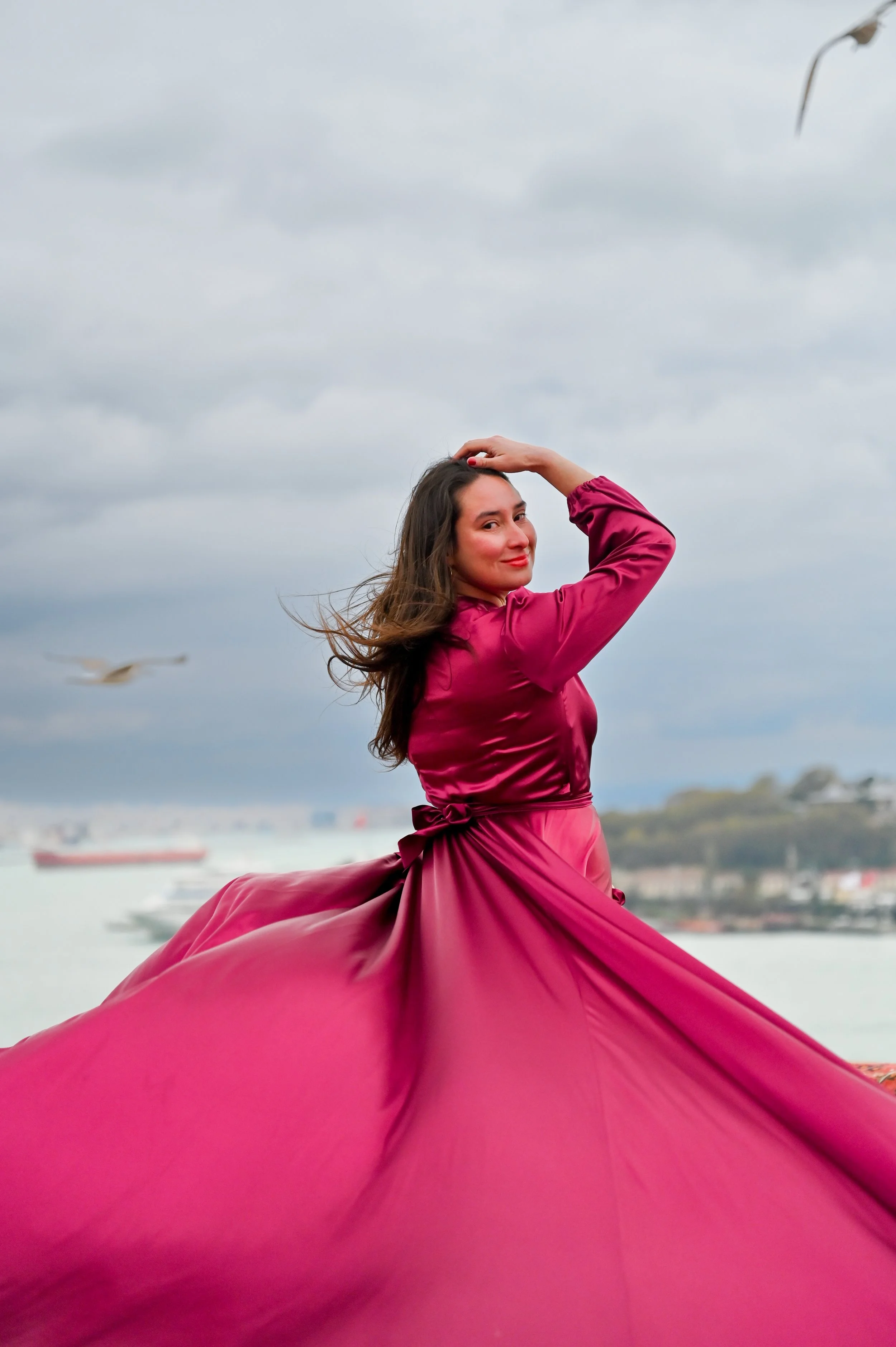 A woman in a pink dress standing outdoors near a body of water with ships in the background, and clouds overhead.