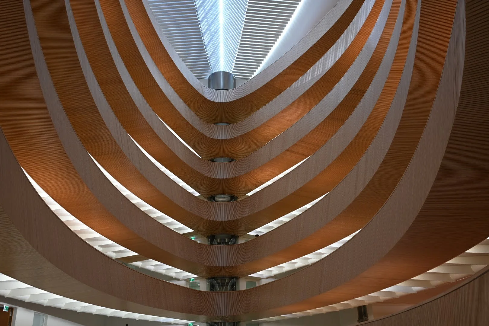 Multiple floors of a modern building viewed from below, with curved wooden balconies and white ceiling panels, illuminated by bright overhead lighting.