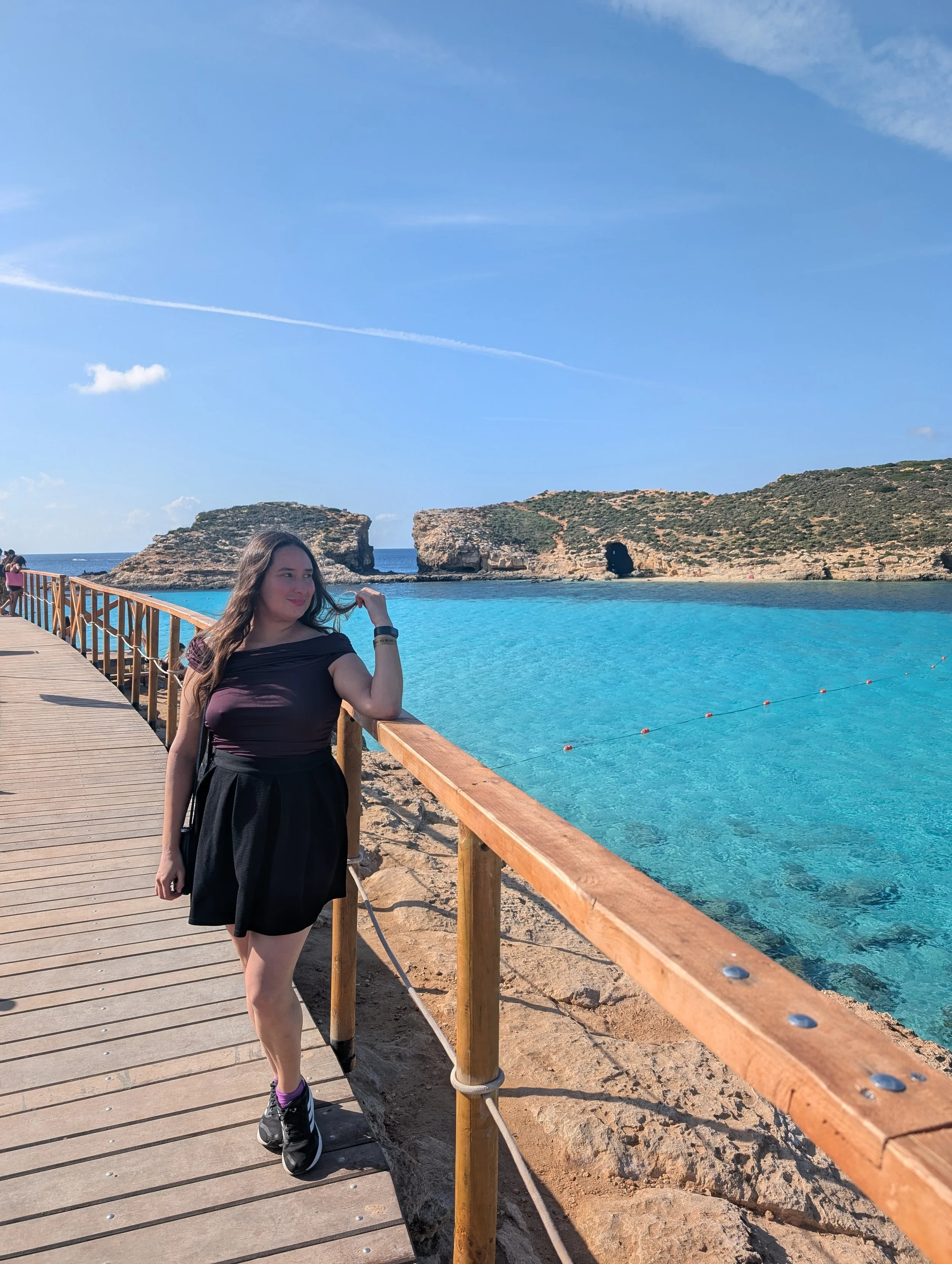 A woman standing on a wooden pier by the ocean, with rocky cliffs and a natural arch in the background under a blue sky with some clouds.