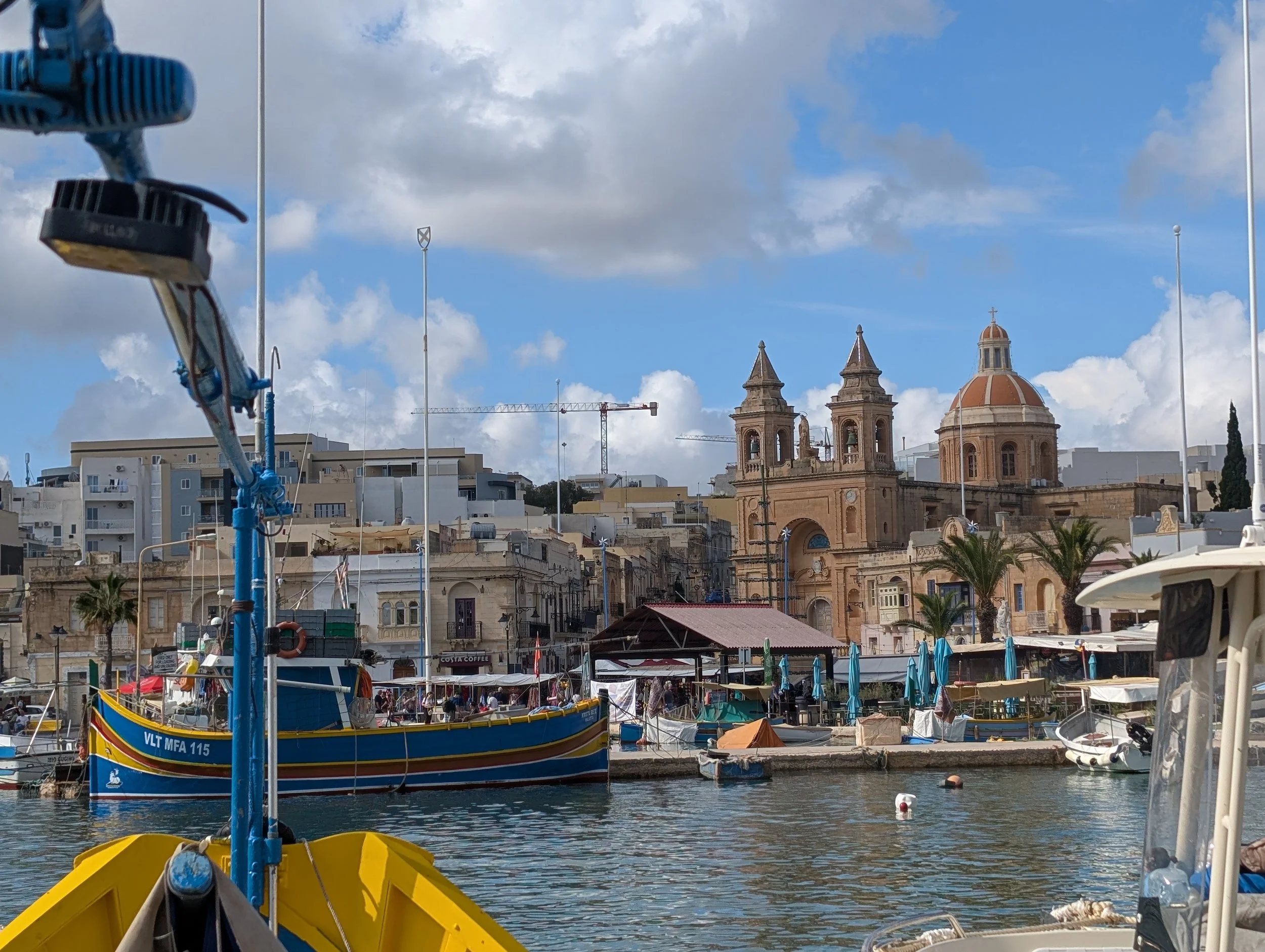 A harbor with boats docked, a historic church with domed and towered architecture, and city buildings in the background under a partly cloudy sky.