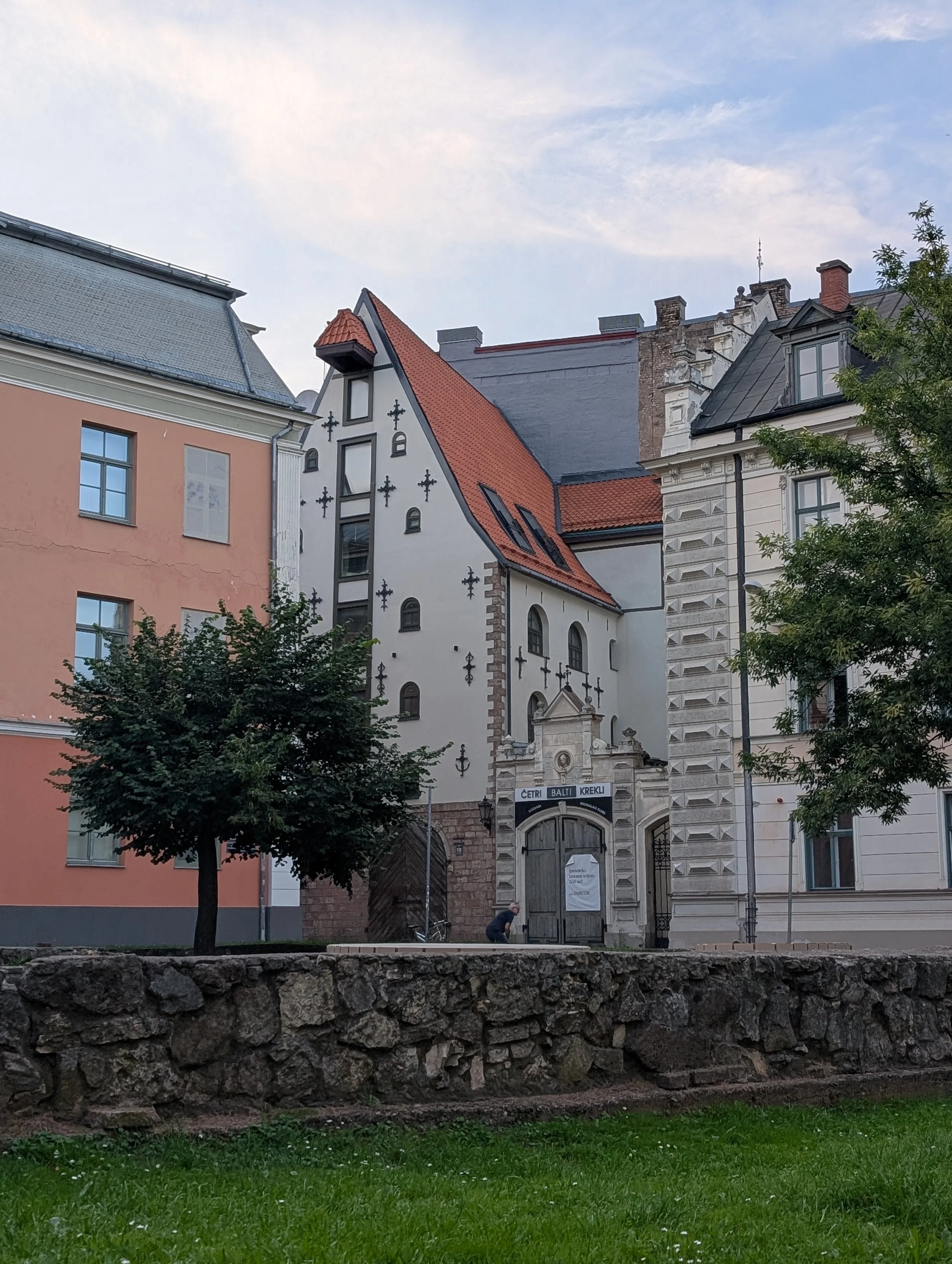 View of historic buildings with different architectural styles, one has mock cross adornments, and there is a stone wall and green lawn in the foreground.