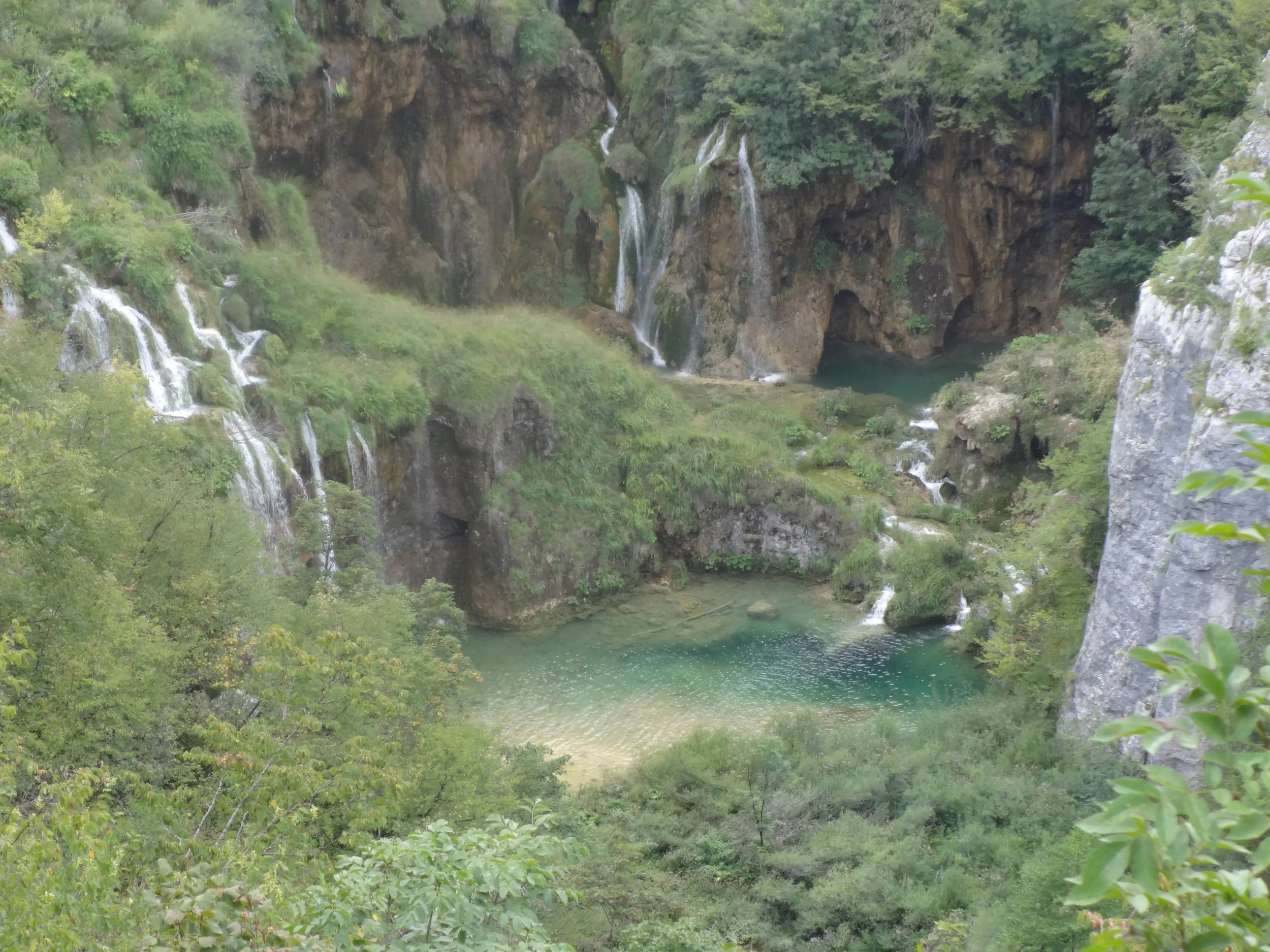 A scenic view of a small waterfall cascading down a rocky hillside into a greenish blue lake surrounded by dense trees and vegetation.