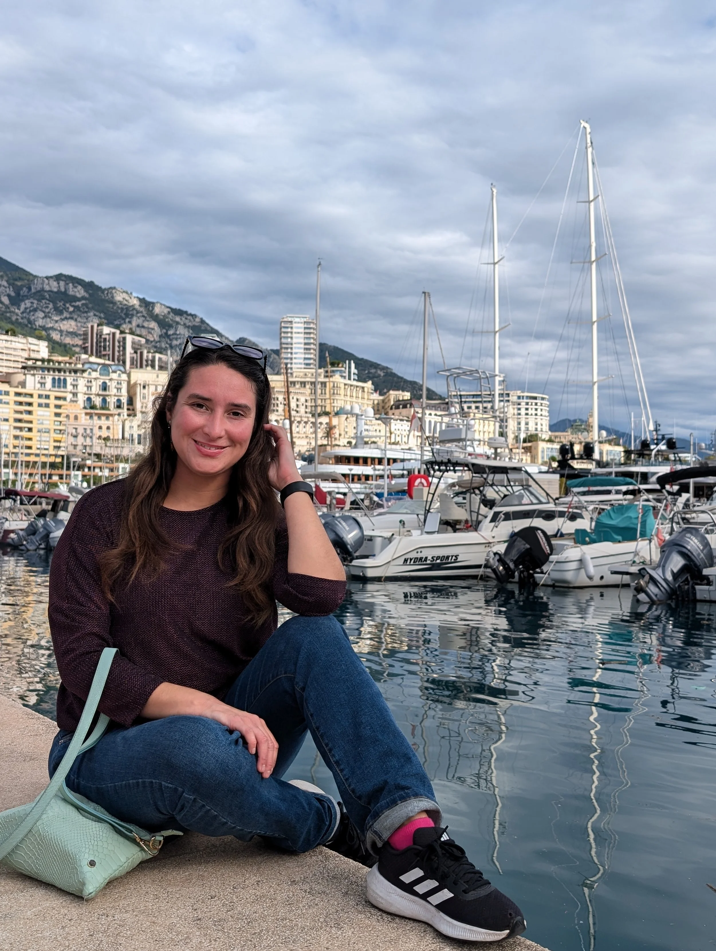 A woman sitting by a marina with boats, buildings, and mountains in the background, wearing a brown sweater, jeans, black sneakers, and sunglasses on her head, with a light green purse beside her.