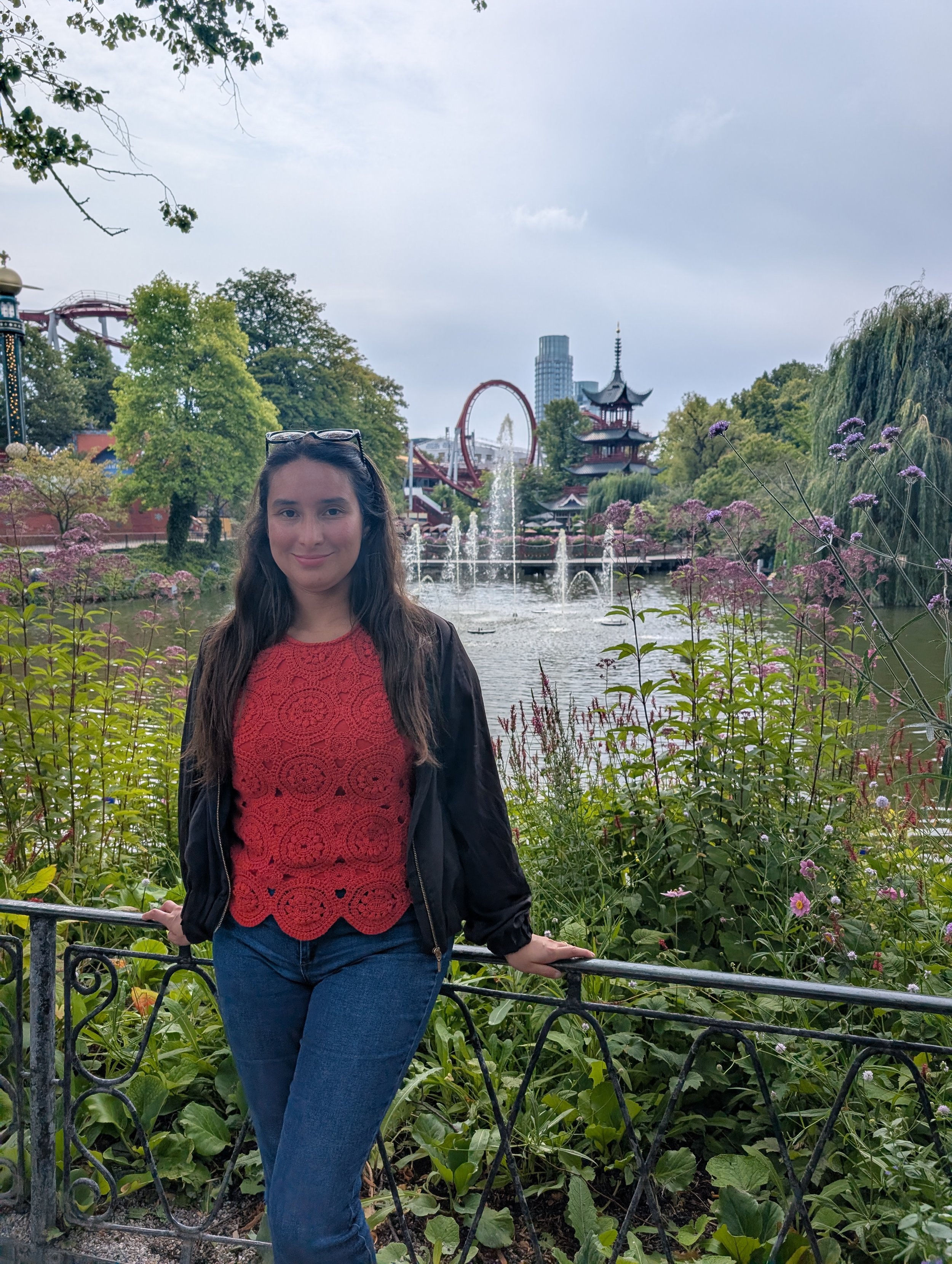 A woman with long dark hair wearing a red lace top and dark jacket, posing in front of a scenic park with water fountains, a Japanese-style pagoda, lush green trees, and a roller coaster in the background.