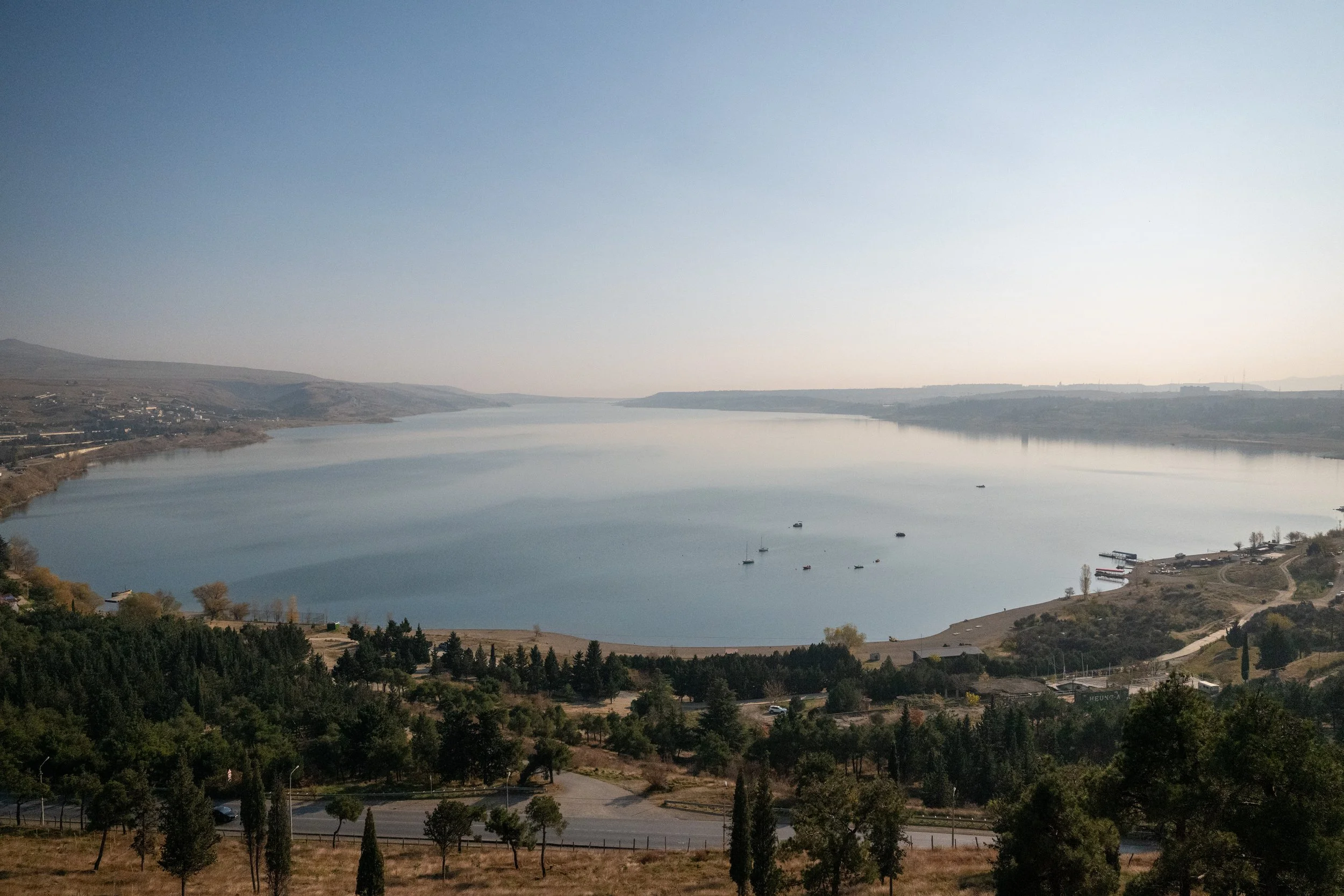 A large river or reservoir with boats docked along the shore, surrounded by rolling hills and trees, under a clear sky.