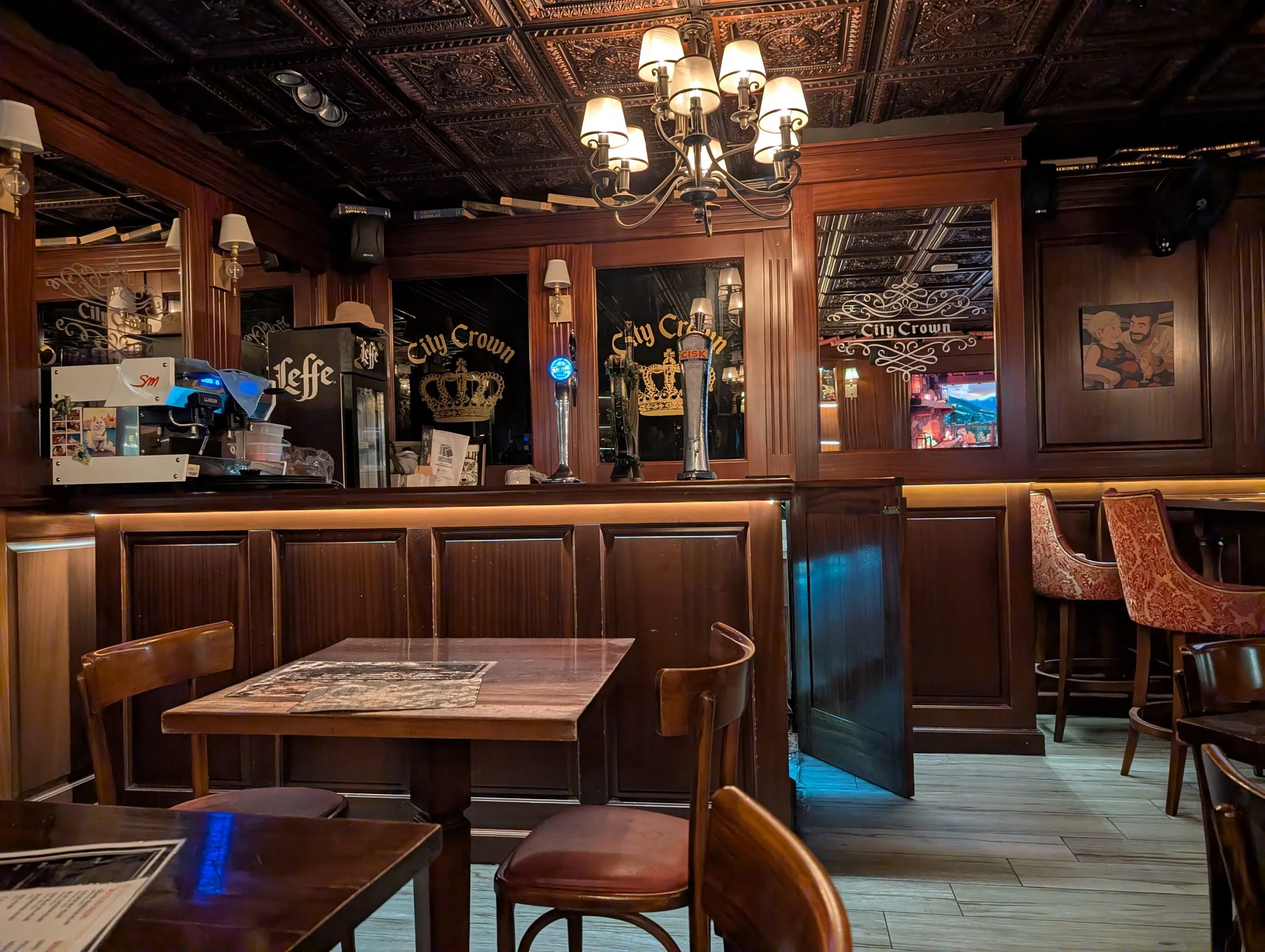 Interior of a cosy restaurant with wooden panelling, a dark, ornate ceiling, and warm lighting. There are tables and chairs, and a bar area with a coffee machine and decorations, including a framed painting on the wall.