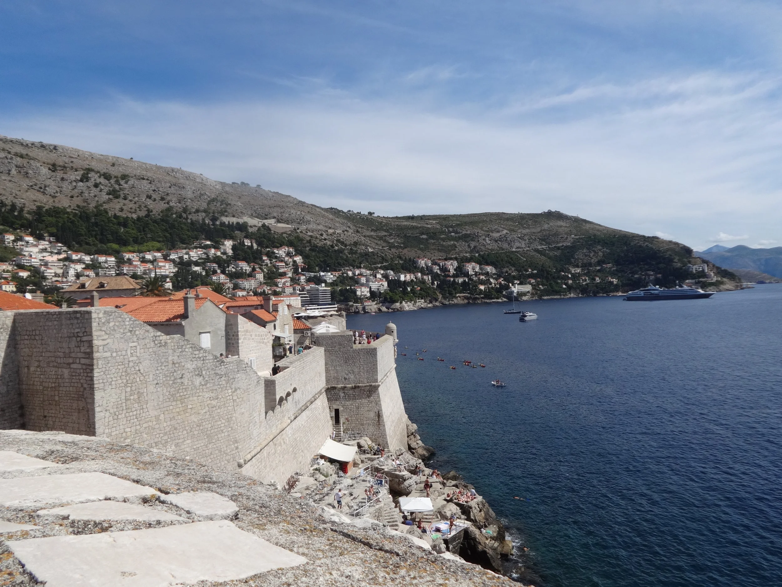 View of a coastal Mediterranean town with stone walls, white buildings with red-tiled roofs, and boats on a calm blue sea, with mountains in the background under a partly cloudy sky.