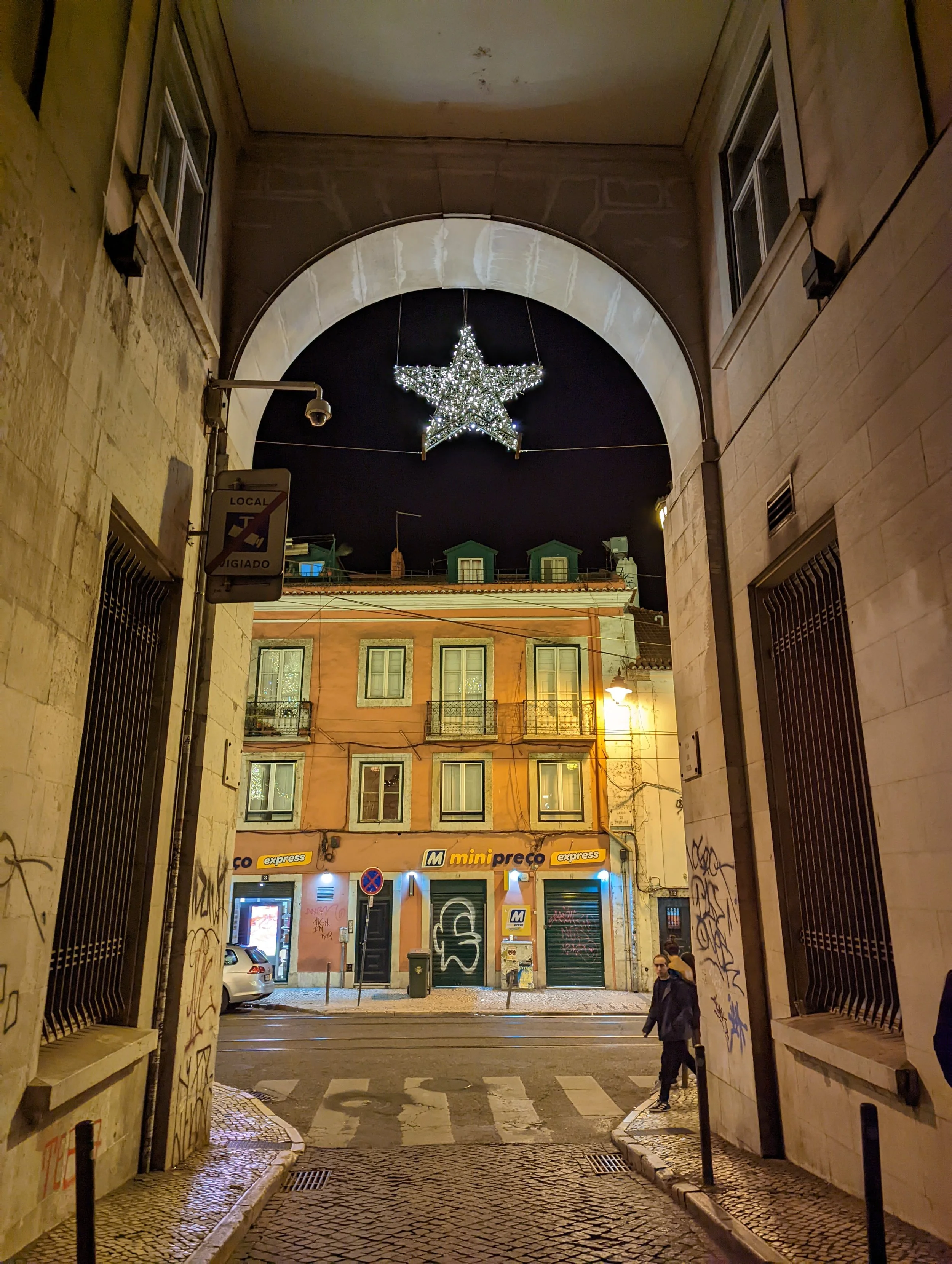 Nighttime street scene viewed through an archway with a decorated illuminated star hanging overhead. Building facades have windows and graffiti, with cars parked and two people walking nearby.
