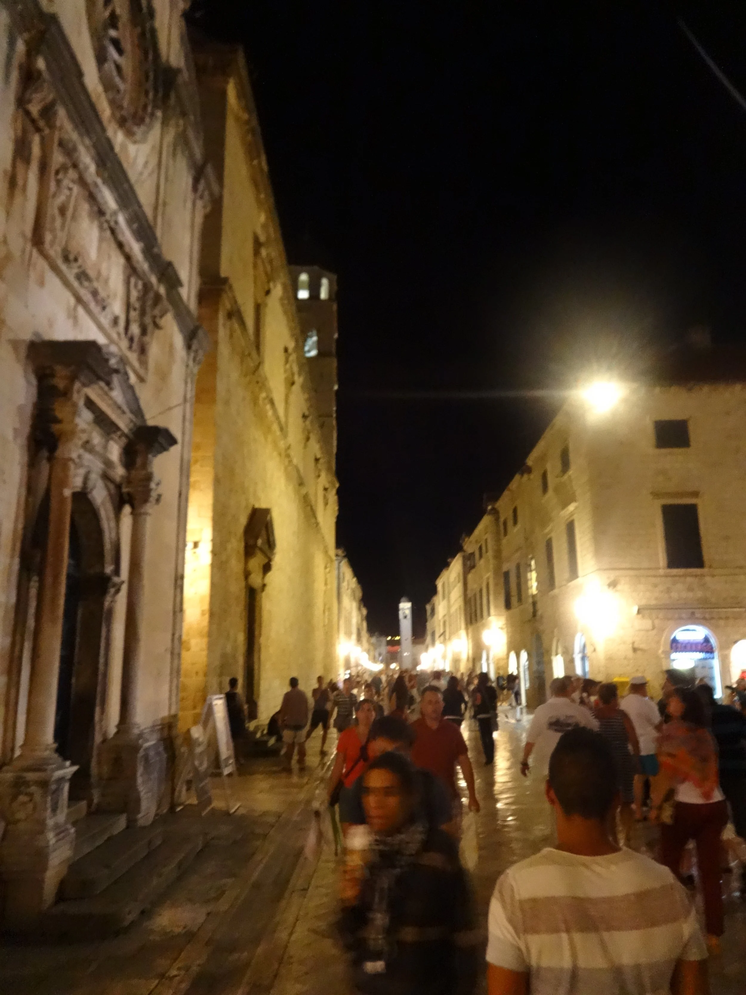 Night scene of a crowded historic street with people walking and sitting at outdoor tables, illuminated by warm streetlights, with old stone buildings on either side.