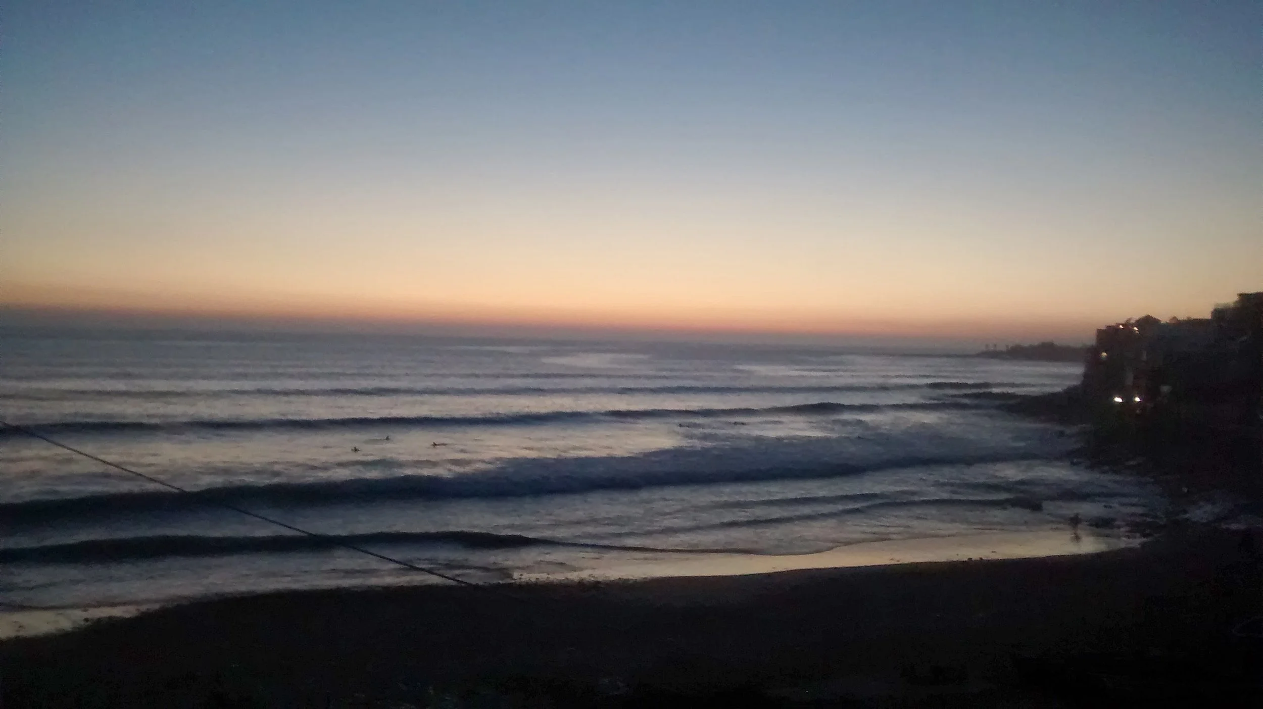 Sunset over a calm ocean with gentle waves, a few surfers in the water, and a shoreline with some buildings and trees in the distance.