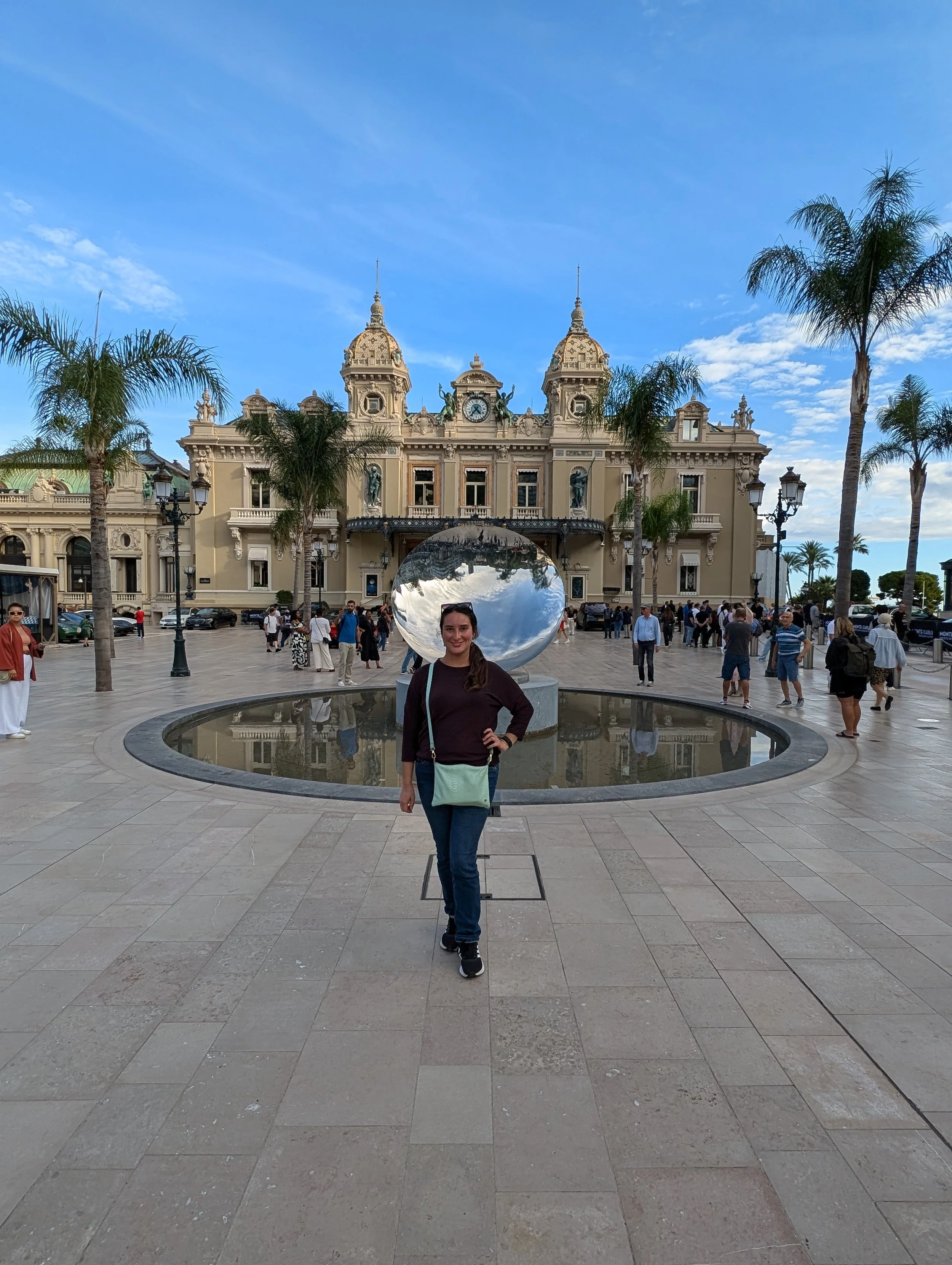 A woman smiling and standing in front of a reflective sculpture in a public square, with an ornate building with towers and a clock in the background, and surrounded by palm trees and other people.