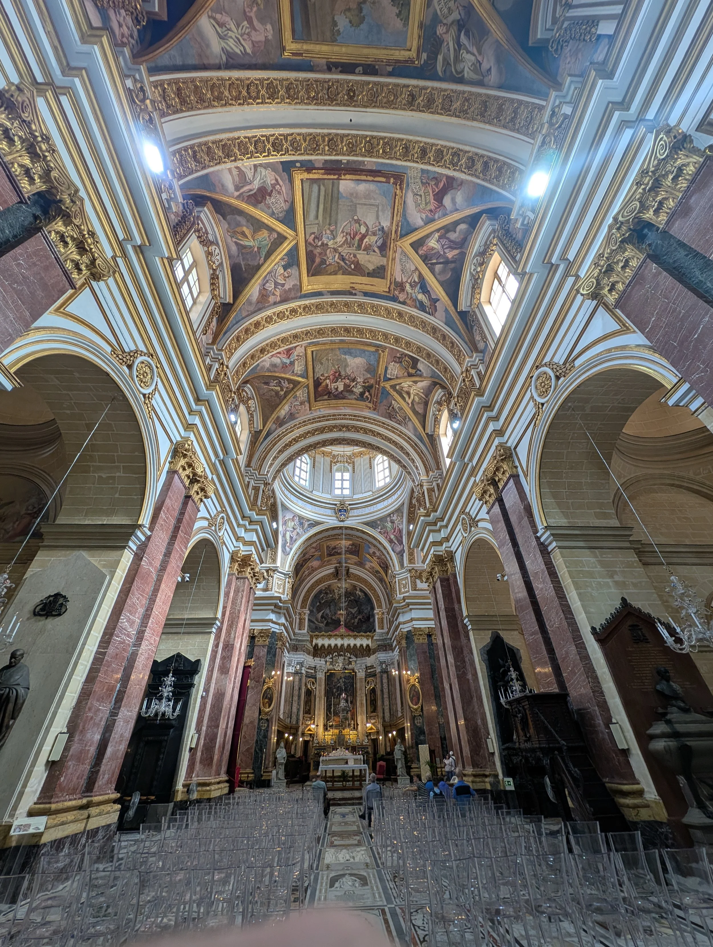 Inside of a grand church or cathedral with a high, ornate ceiling decorated with colorful religious paintings and gold accents. There are tall windows letting in natural light, and an altar area at the far end with religious statues and artwork. Seve