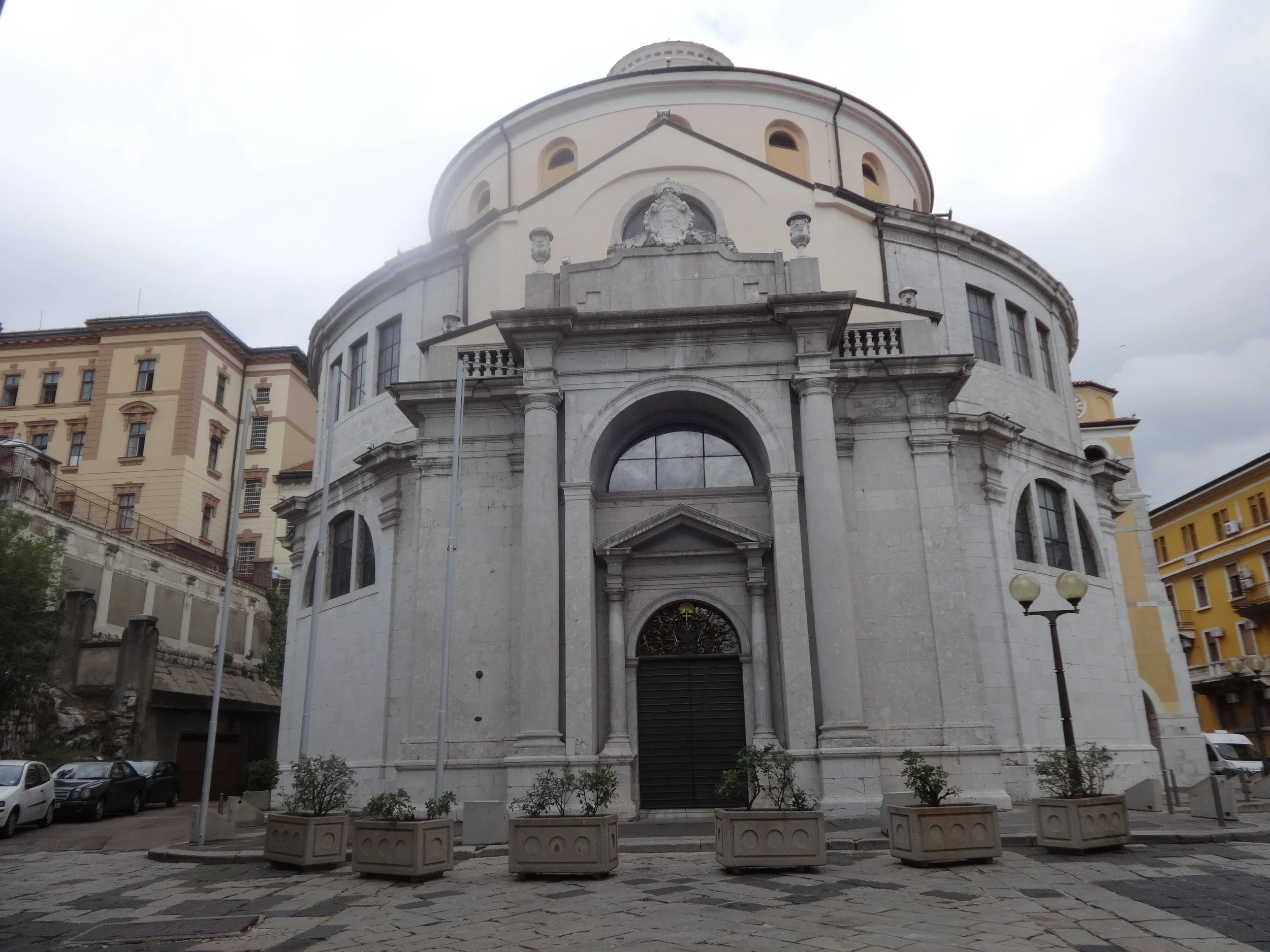 Front view of a historic church with gray stone facade, large arched entrance, and a rounded dome, surrounded by parked cars and city buildings under a cloudy sky.