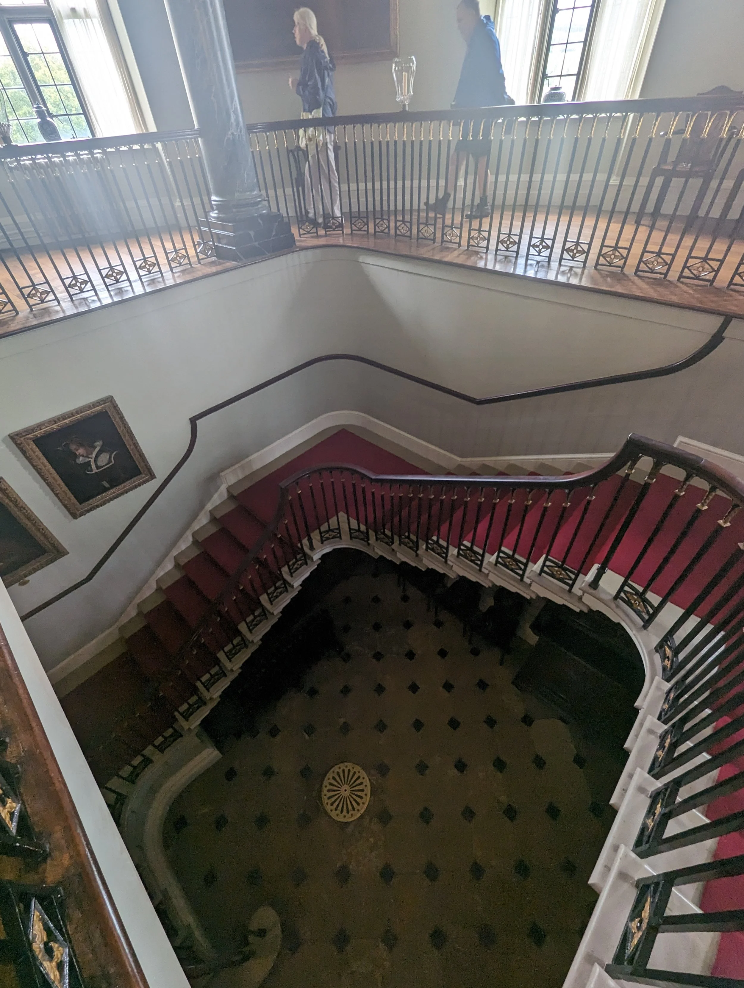Interior view of a staircase looking down from the second floor, with a decorative wooden railing and black and tan patterned tile flooring at the bottom. The staircase has red carpeted steps and black and gold decorative elements on the railing. The