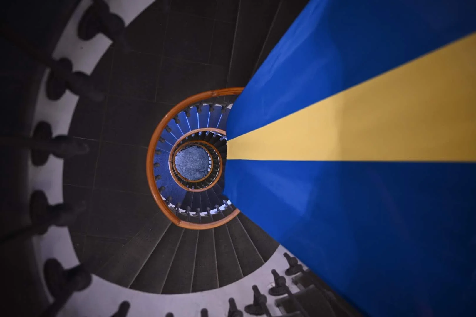 A spiral staircase viewed from above, with a blue and yellow cloth hanging down the center. The staircase has black steps and a wooden handrail.