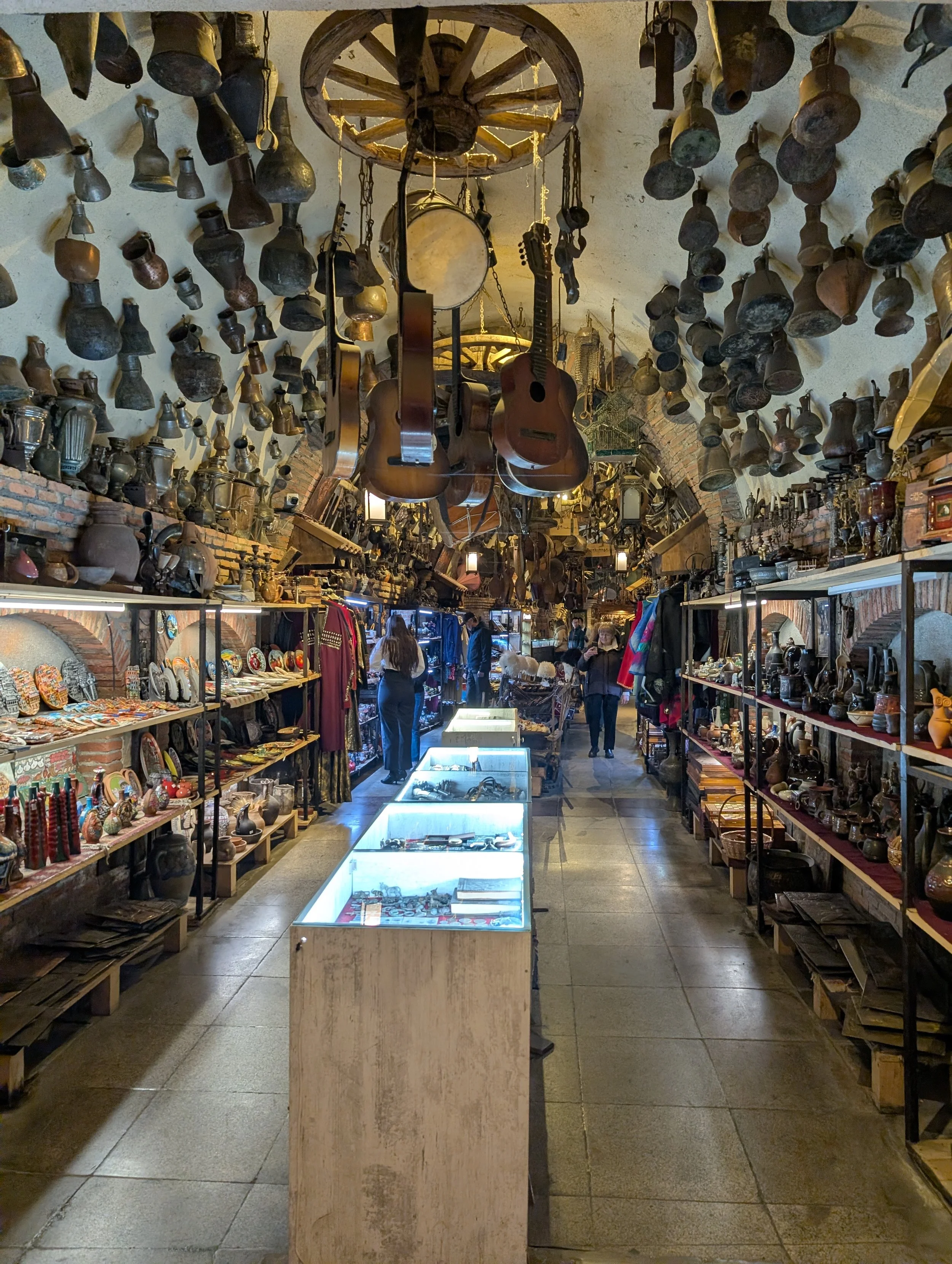 Interior of a vintage shop with hanging old pots, clocks, and musical instruments, including guitars, with shelves displaying various collectibles and artifacts, and people browsing.