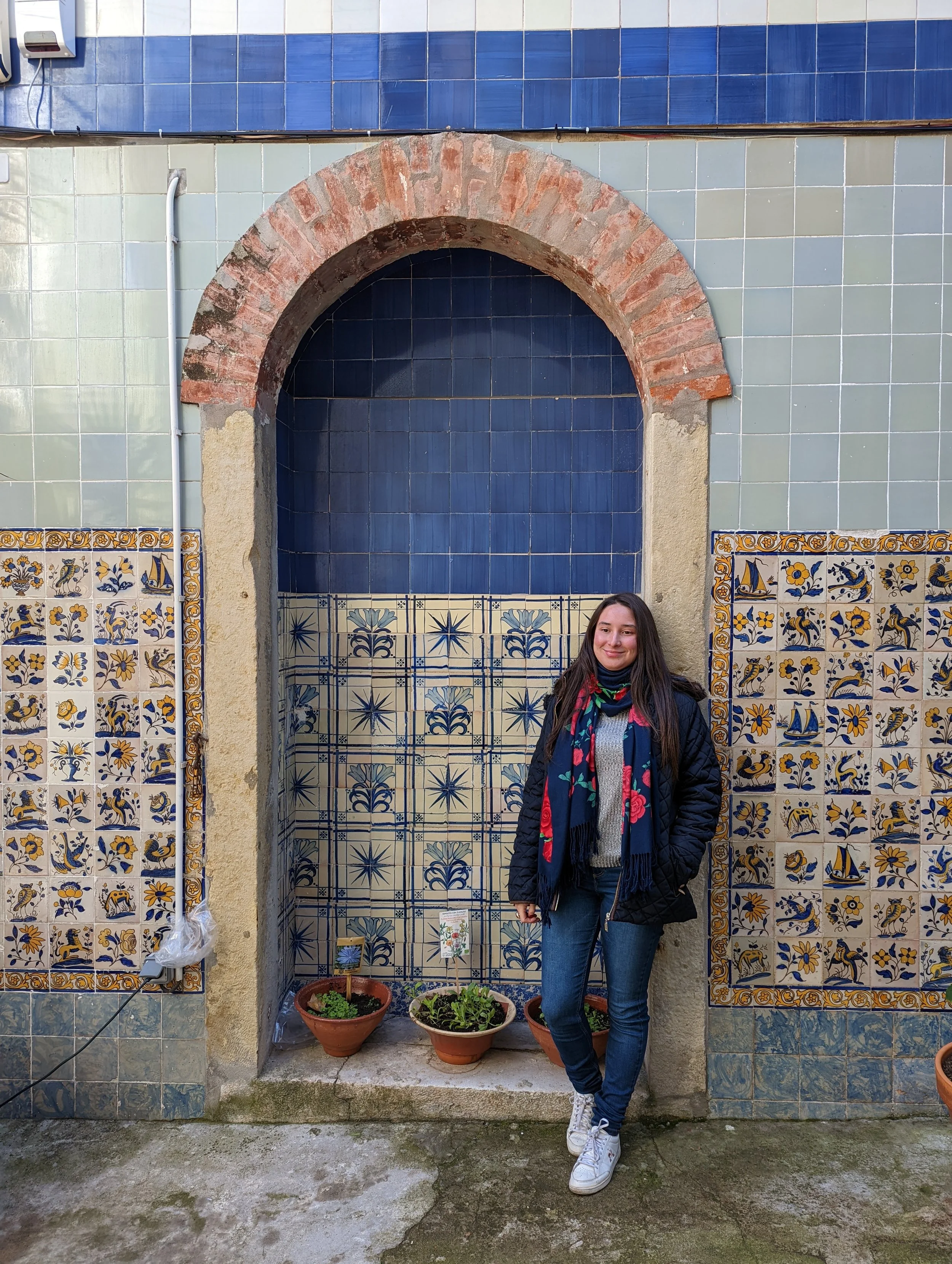 Young woman standing in front of a decorative tiled wall with arrows, sailboats, birds, and floral designs. She wears a black jacket, patterned gray sweater, blue jeans, white sneakers, and a colorful scarf. There are potted plants on the ground and 