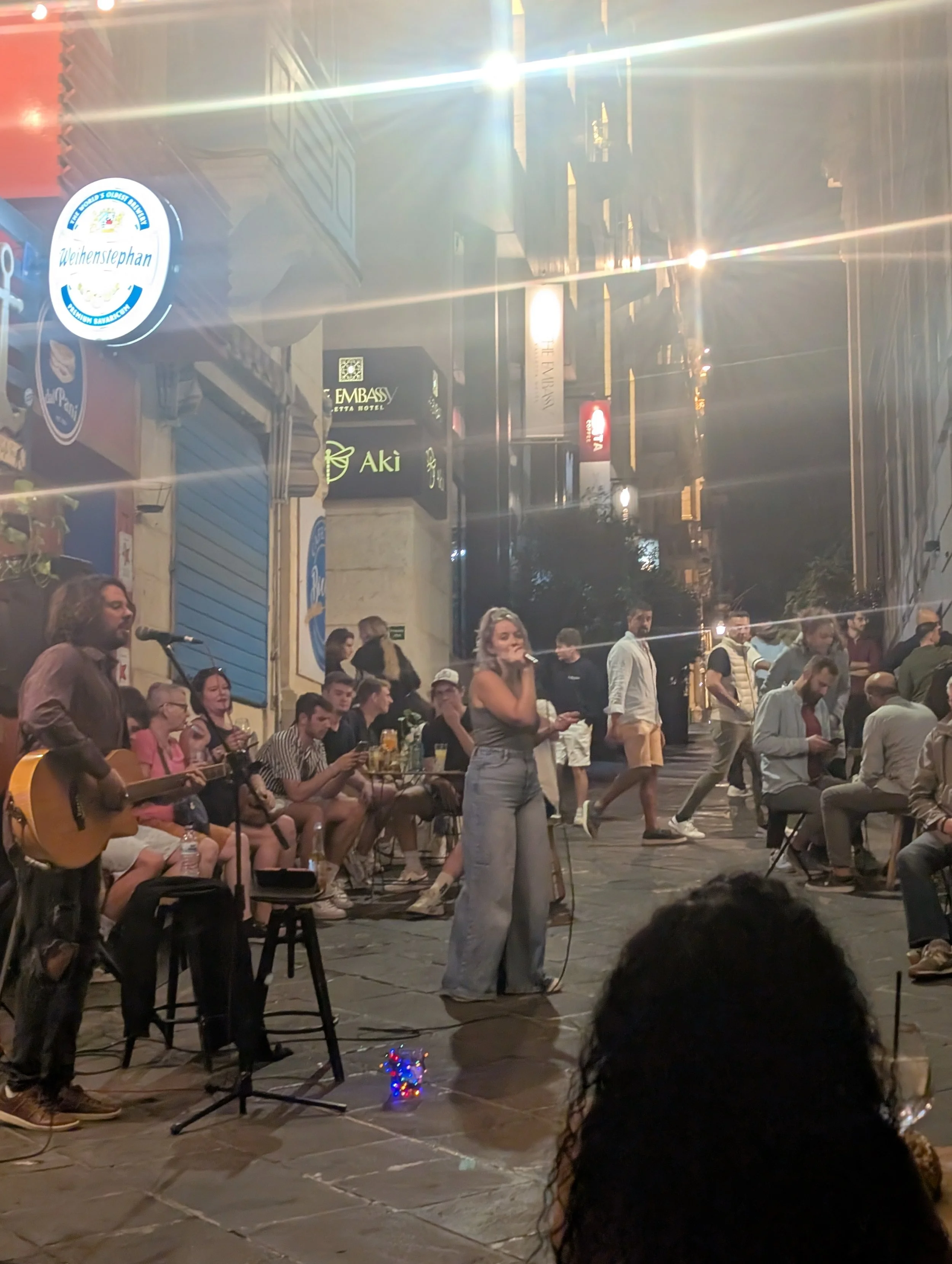 Group of people gathered outdoors at night, some seated with drinks, others standing, with a woman singing into a microphone, a guitarist playing, and various signs advertising businesses in the background.