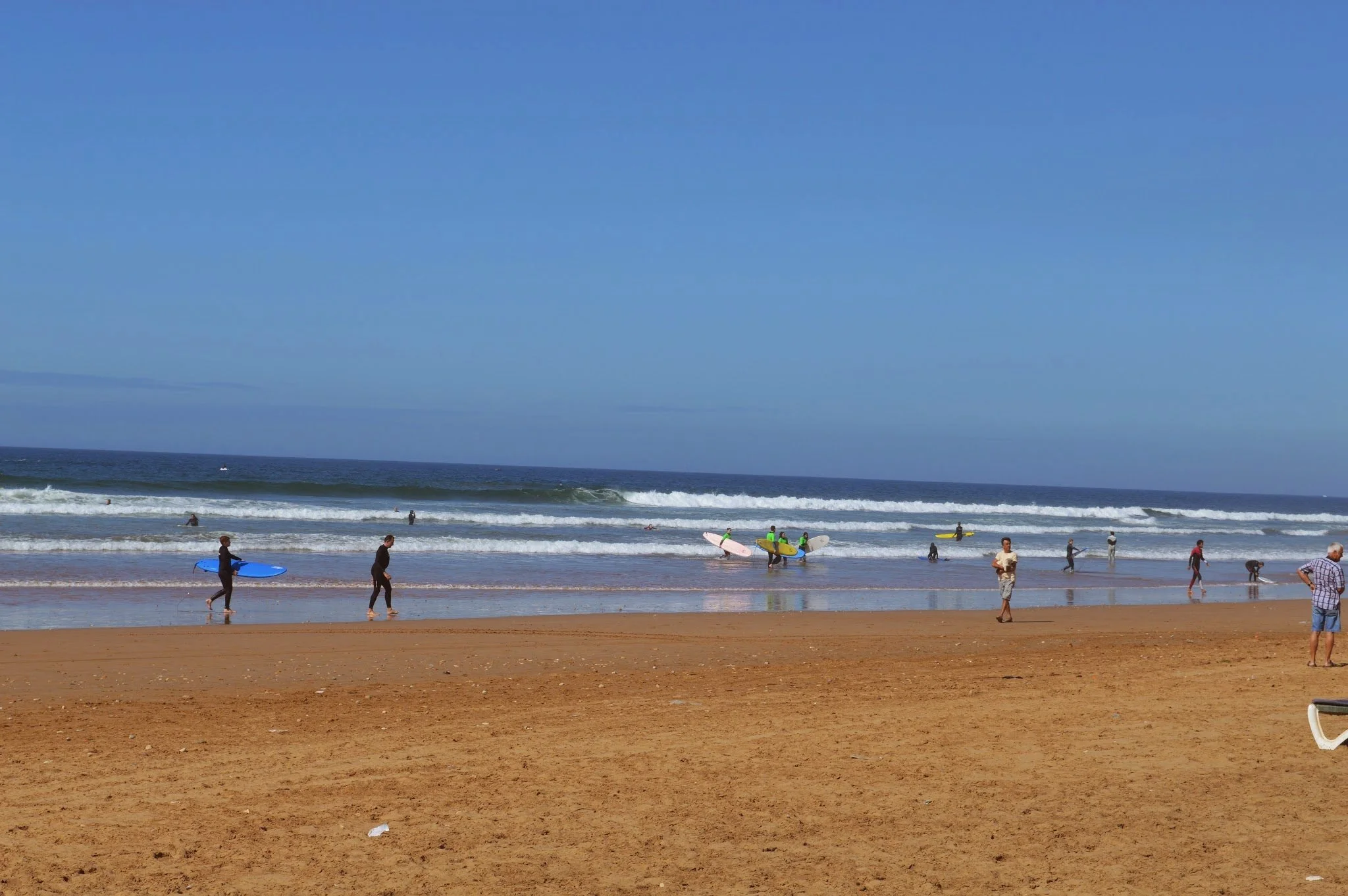 People walking and surfing on the beach with waves in the ocean and a clear blue sky.