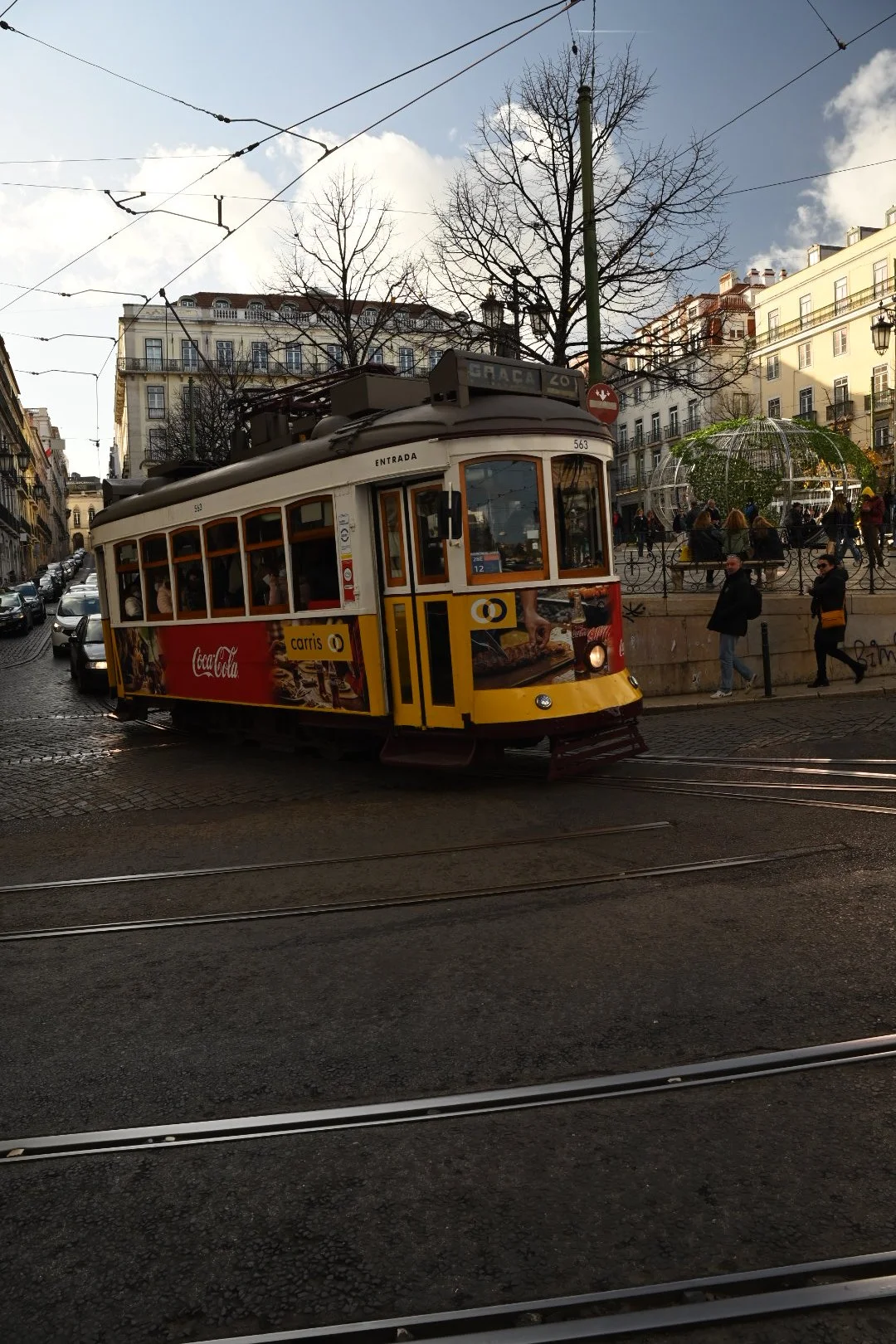 A yellow tram traveling on tracks in a city street with cobblestone pavement, surrounded by buildings, leafless trees, and pedestrians. An advertisement for Coca-Cola is visible on the tram.