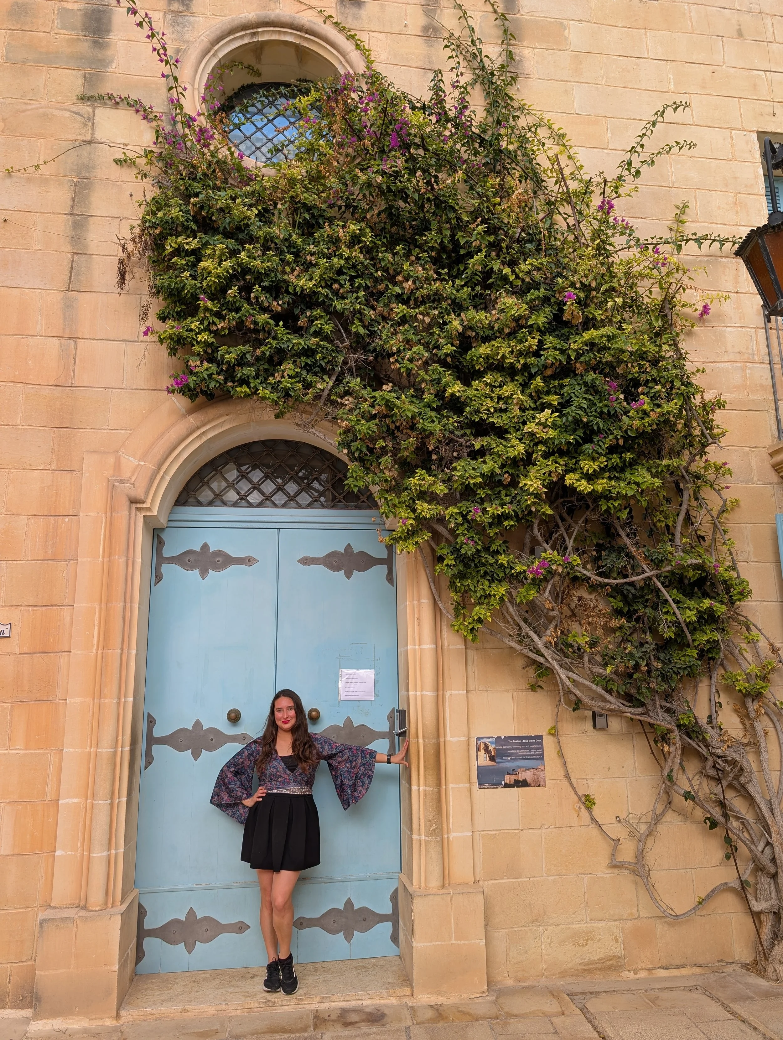 A woman in a flowy patterned top and black skirt standing with one arm extended, touching a large blue door with black decorative accents, in front of a stone building with ivy and purple flowers.