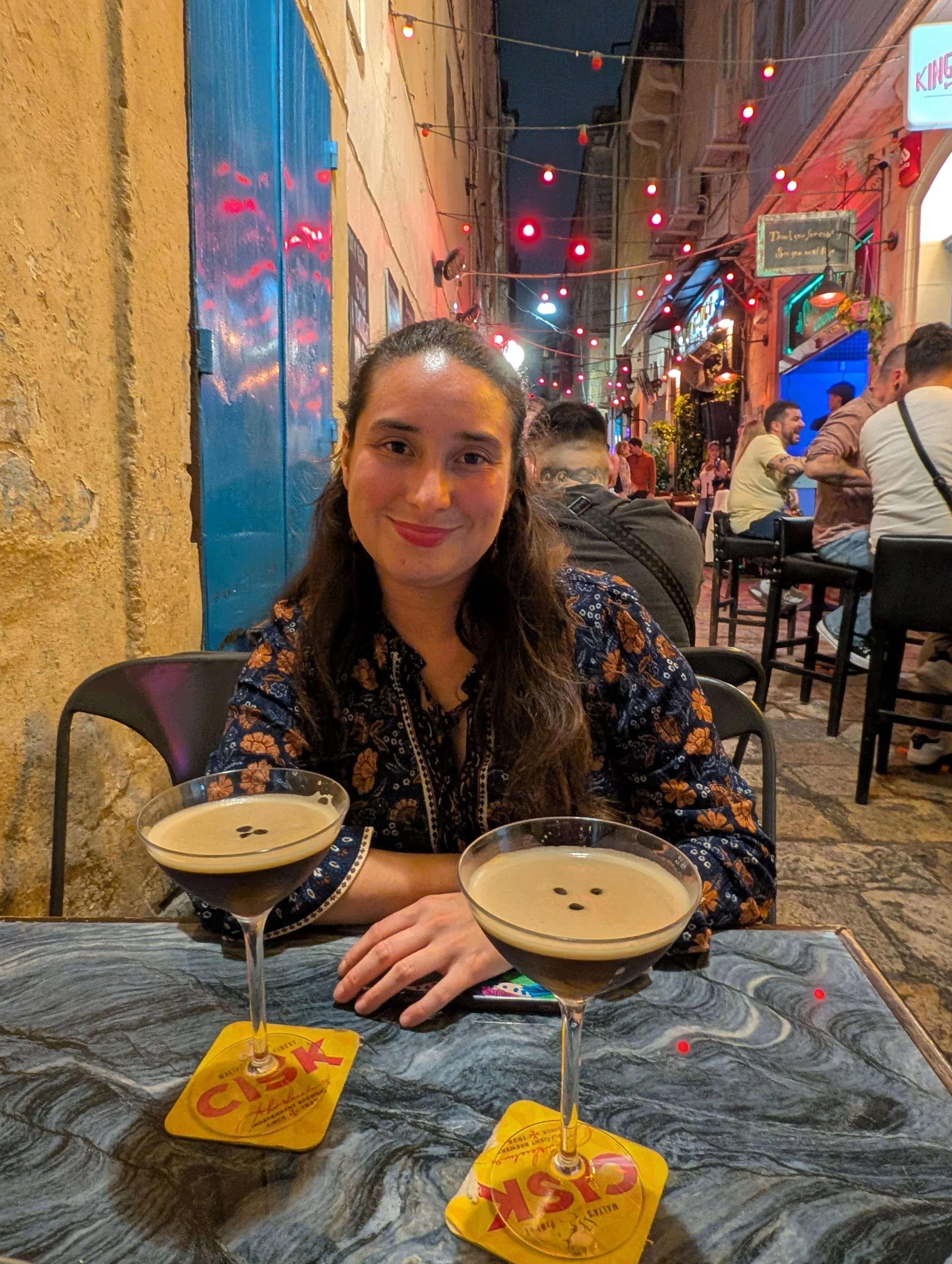 A woman is sitting at a table in an outdoor dining area at night, smiling, with two cocktails in front of her, and other patrons in the background, decorated with string lights.
